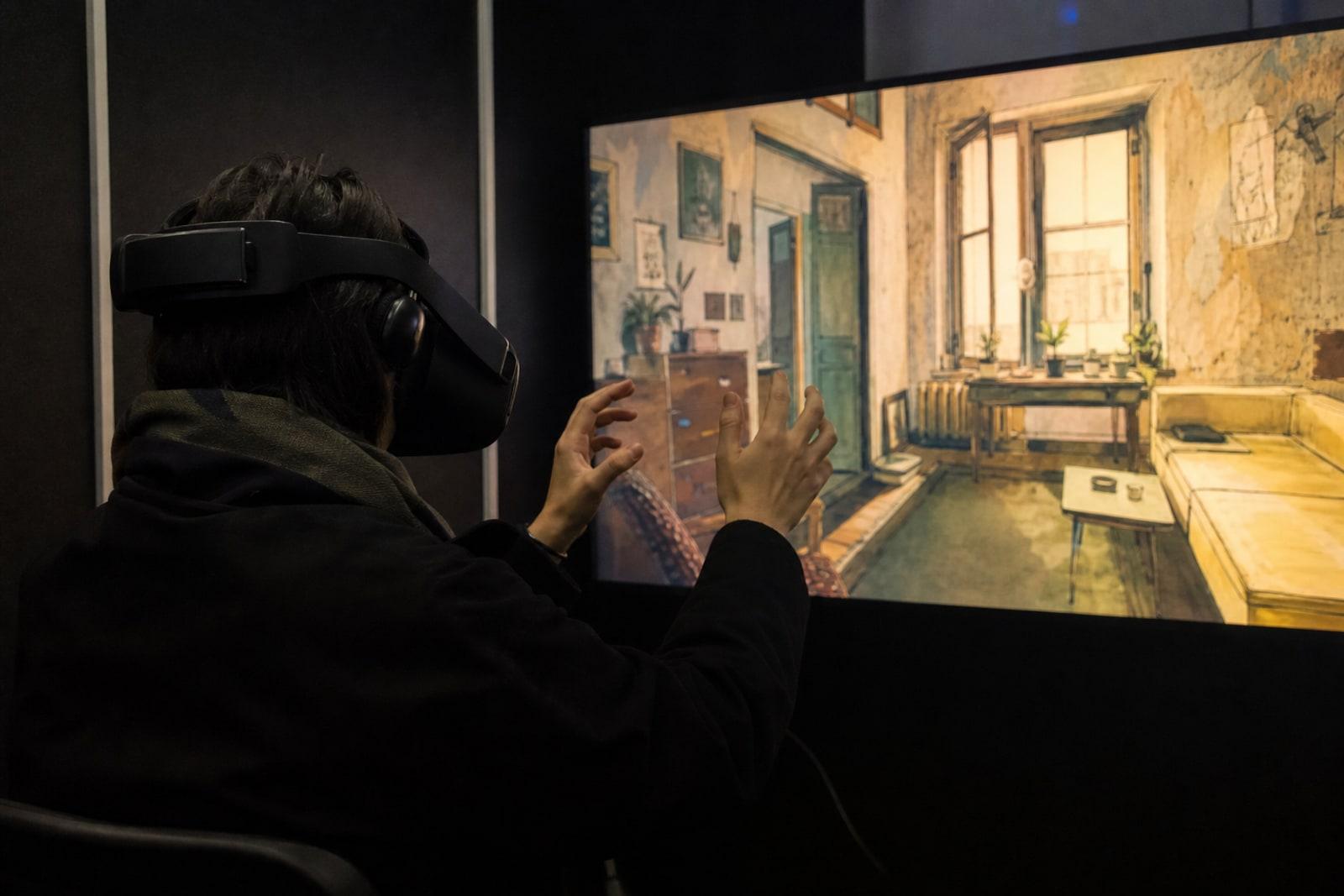 A seated festival visitor wearing a black VR headset in a darkened installation booth, lit by a screen showing a hand-drawn apartment interior.