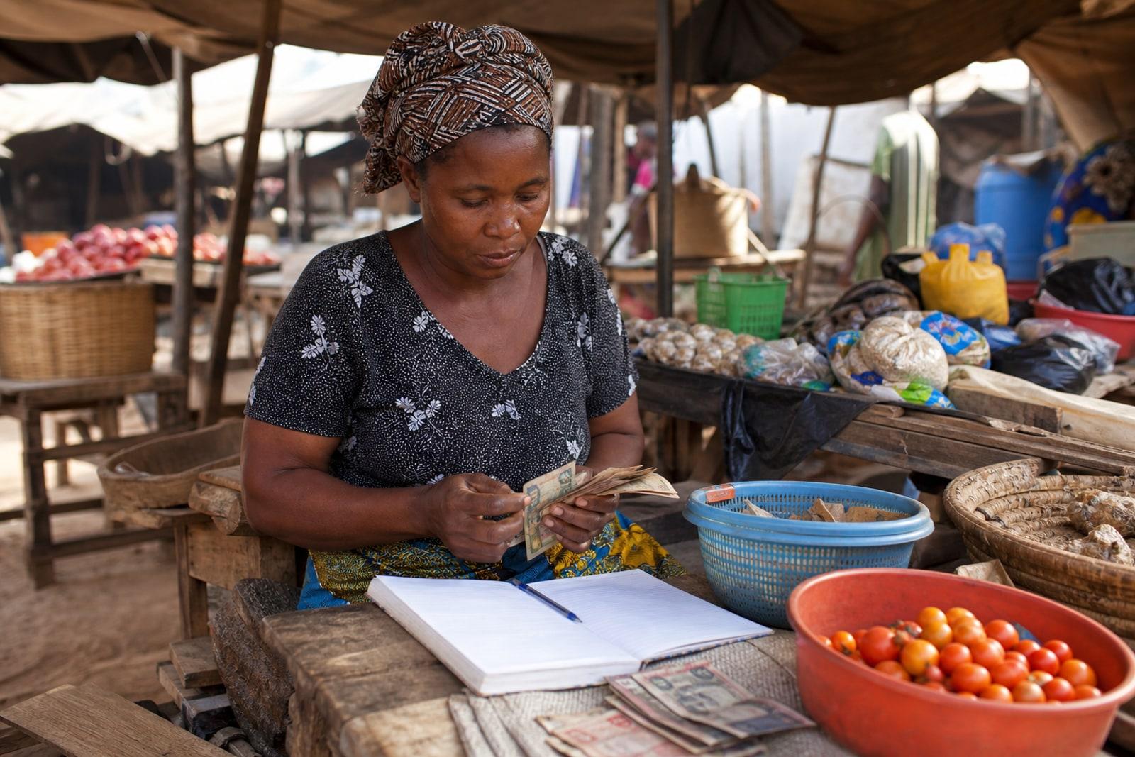 A woman tends a small open-air market stall in Sub-Saharan Africa, counting banknotes beside a paper ledger.