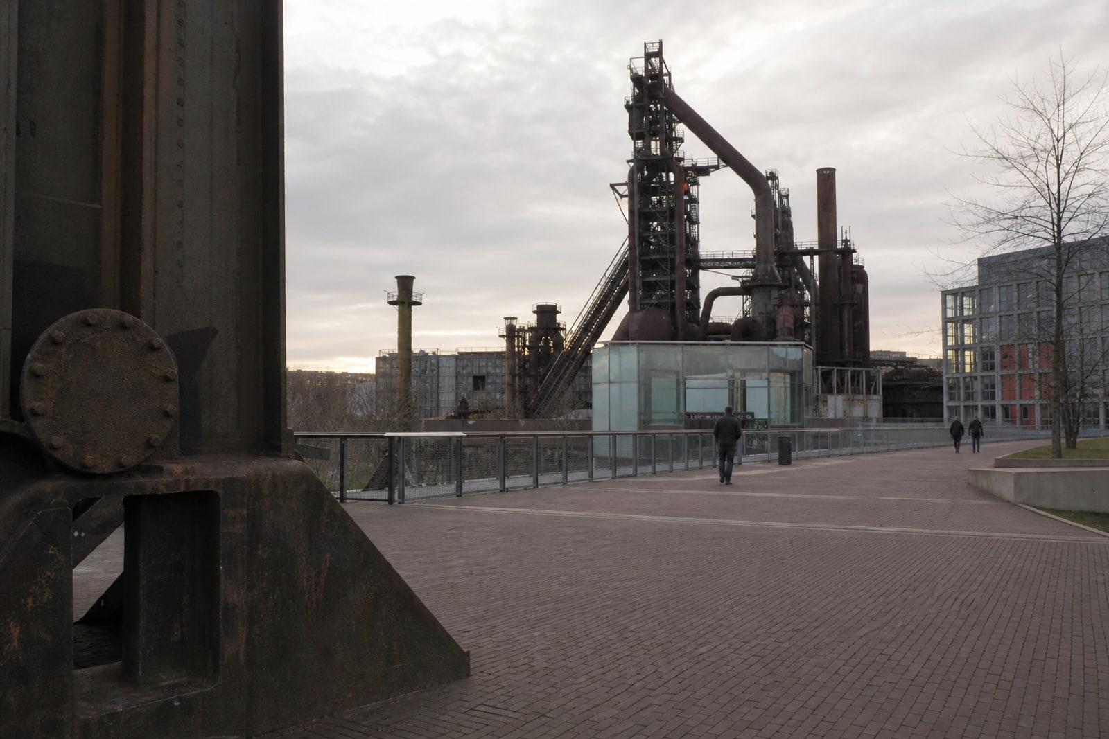 Quiet walkway beside preserved Belval steelworks structures in Esch-sur-Alzette, with anonymous distant pedestrians under overcast natural light.