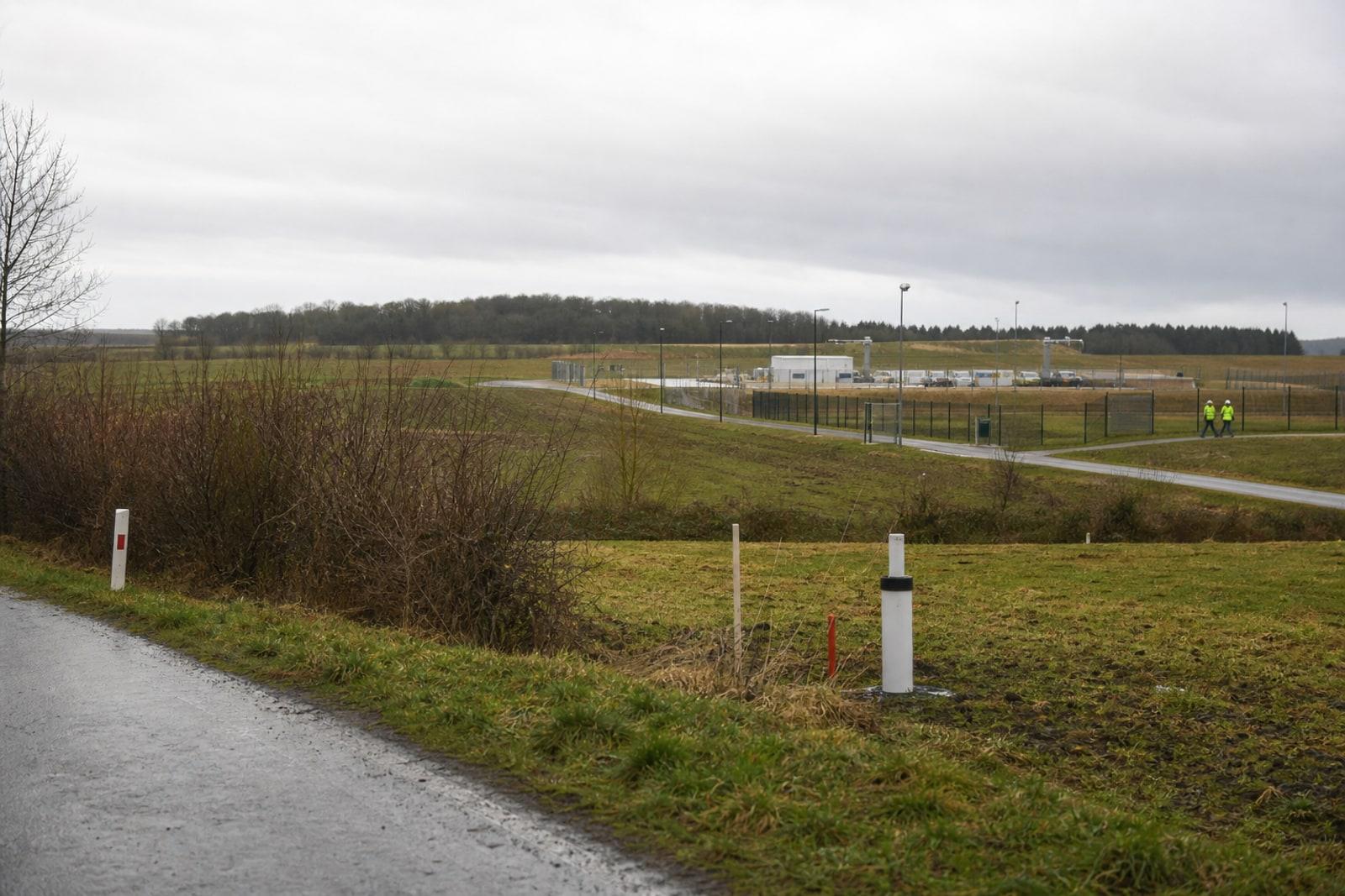 Wet farmland near a fenced rural industrial compound in the Meuse countryside, with distant anonymous workers and monitoring infrastructure under grey daylight.