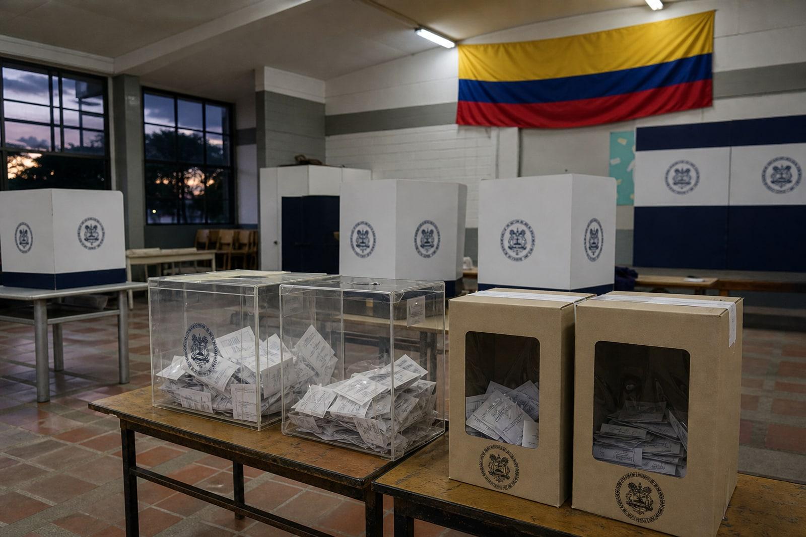 A Colombian polling station with transparent ballot boxes and white voting screens beneath a yellow-blue-red Colombian flag.