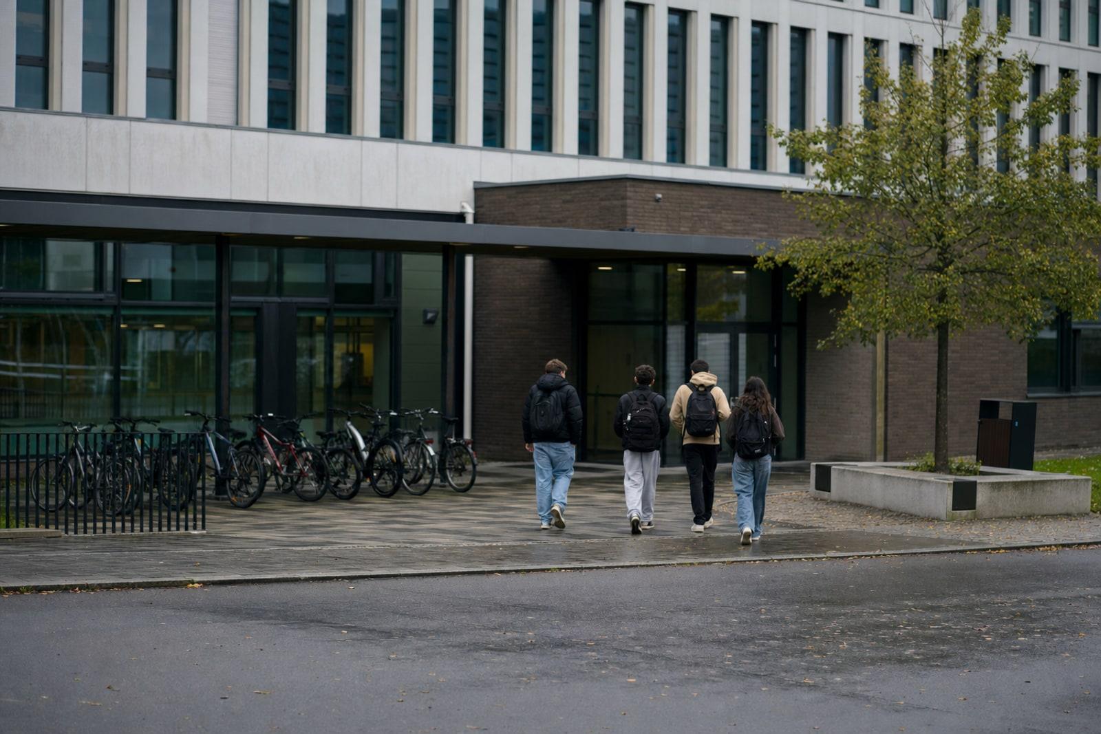 Anonymous teenagers with backpacks walk toward a modern Luxembourg school building on a wet overcast morning.