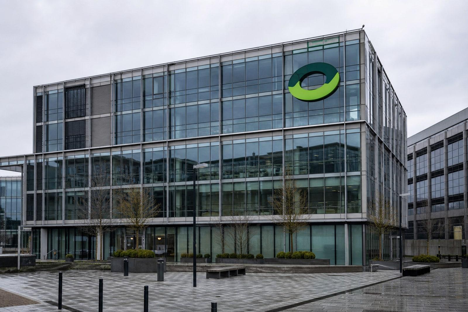 A modern glass-fronted Luxembourg insurance headquarters in the Leudelange office park under grey skies, carrying a green corporate emblem.