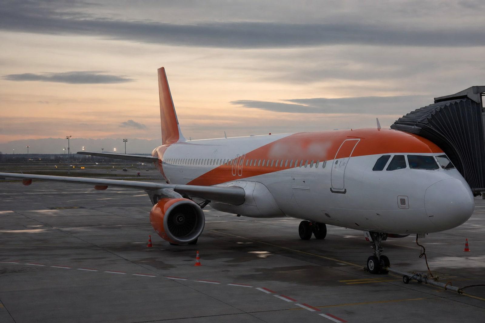 An easyJet Airbus A320 in white livery with bright orange engine cowlings and winglets parked at an airport gate at dusk.