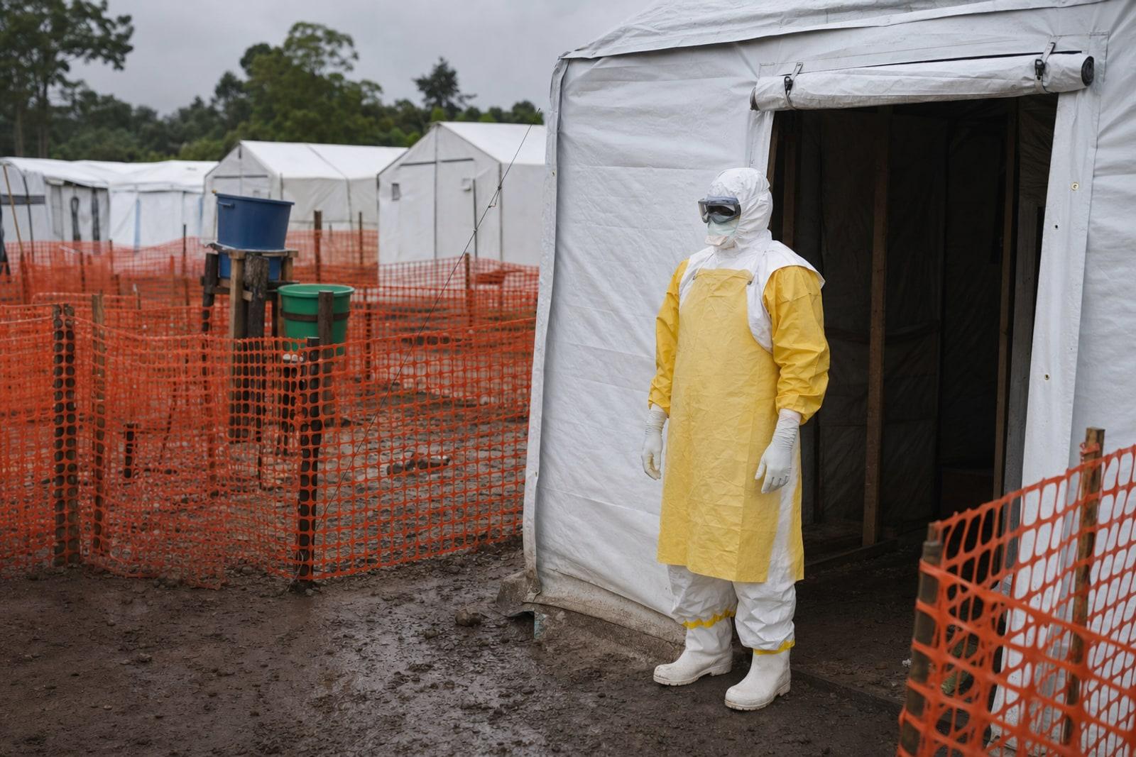 A health worker in full white-and-yellow protective suit, goggles and gloves at the entrance of a tented Ebola treatment centre.
