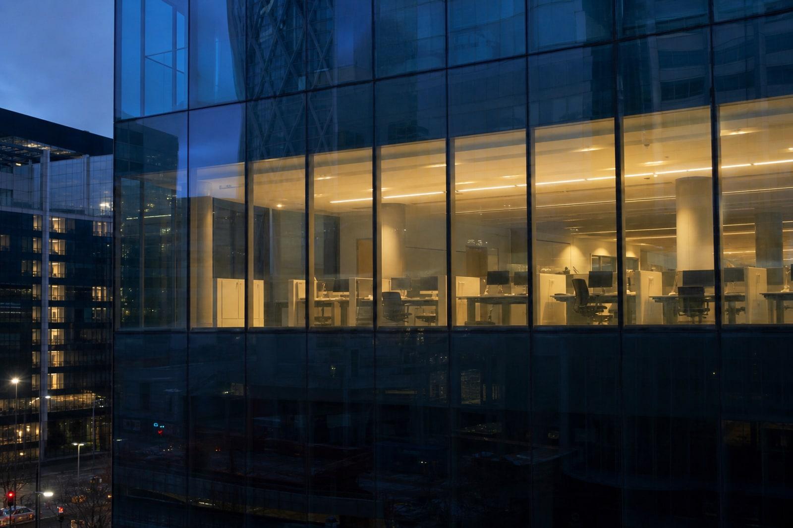 An empty, glass-walled office floor in a financial-district tower, lit from within at dusk and seen from outside.