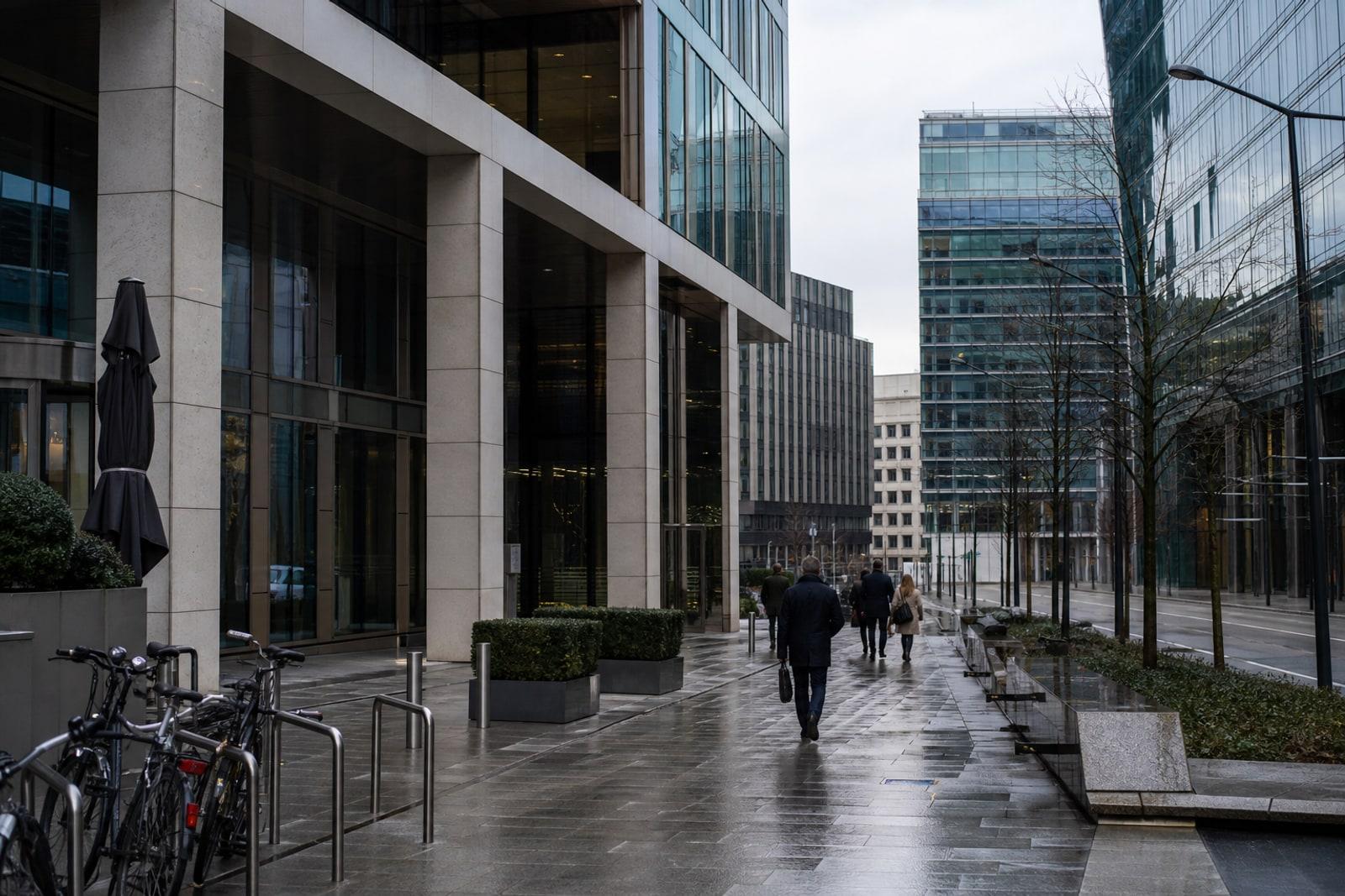 Wet pavement outside modern Luxembourg financial-district office buildings with anonymous commuters walking past glass facades and parked bicycles.