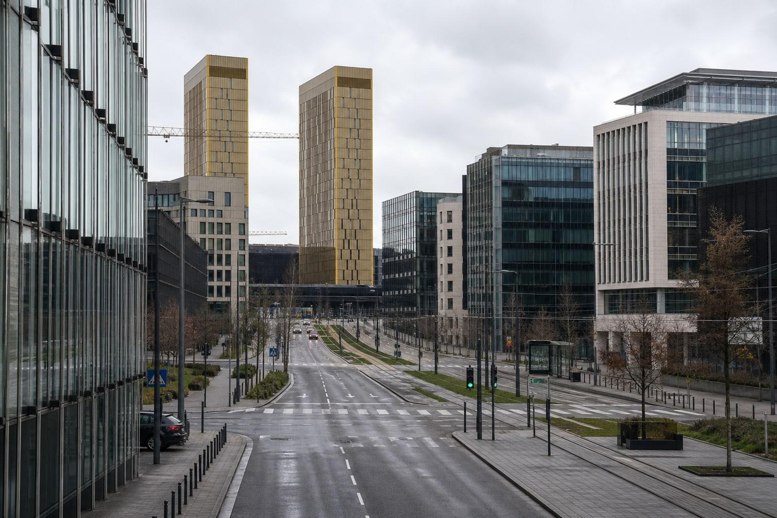 Modern glass-and-steel bank towers on Luxembourg City's Kirchberg plateau, with the gold-bronze twin towers of the EU Court of Justice behind, under an overcast sky.