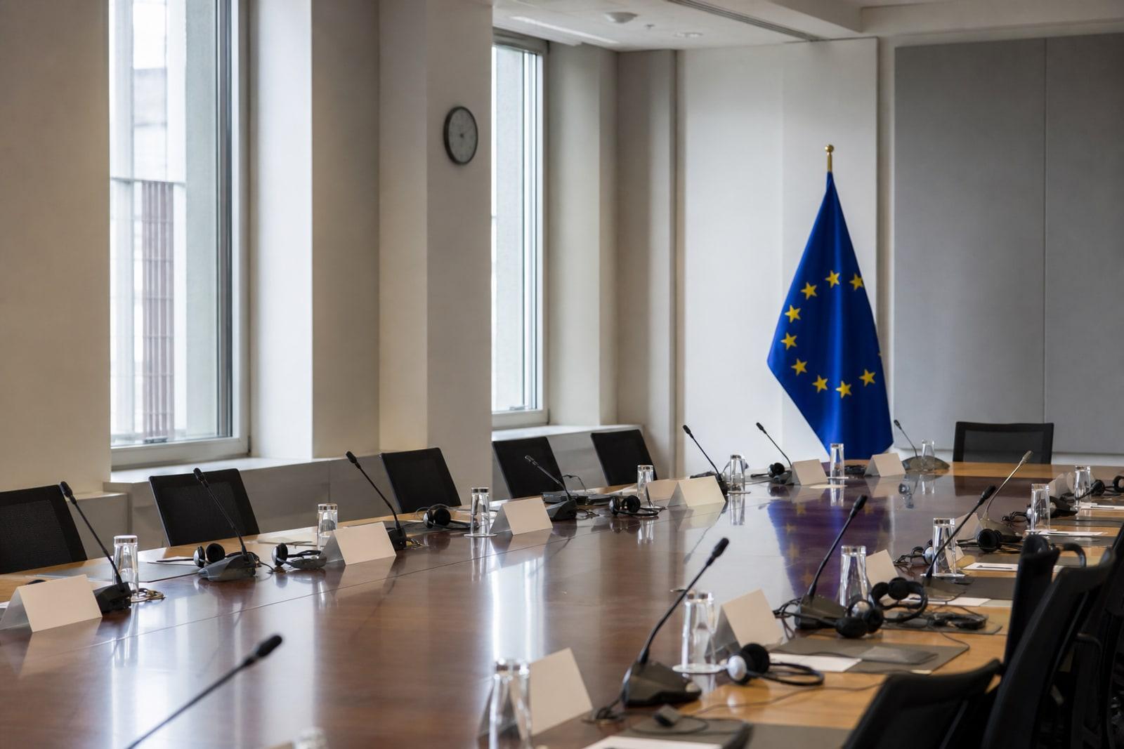An empty closed-door EU conference room in Brussels with a European Union flag beside a table set with name placards and interpreter headsets.