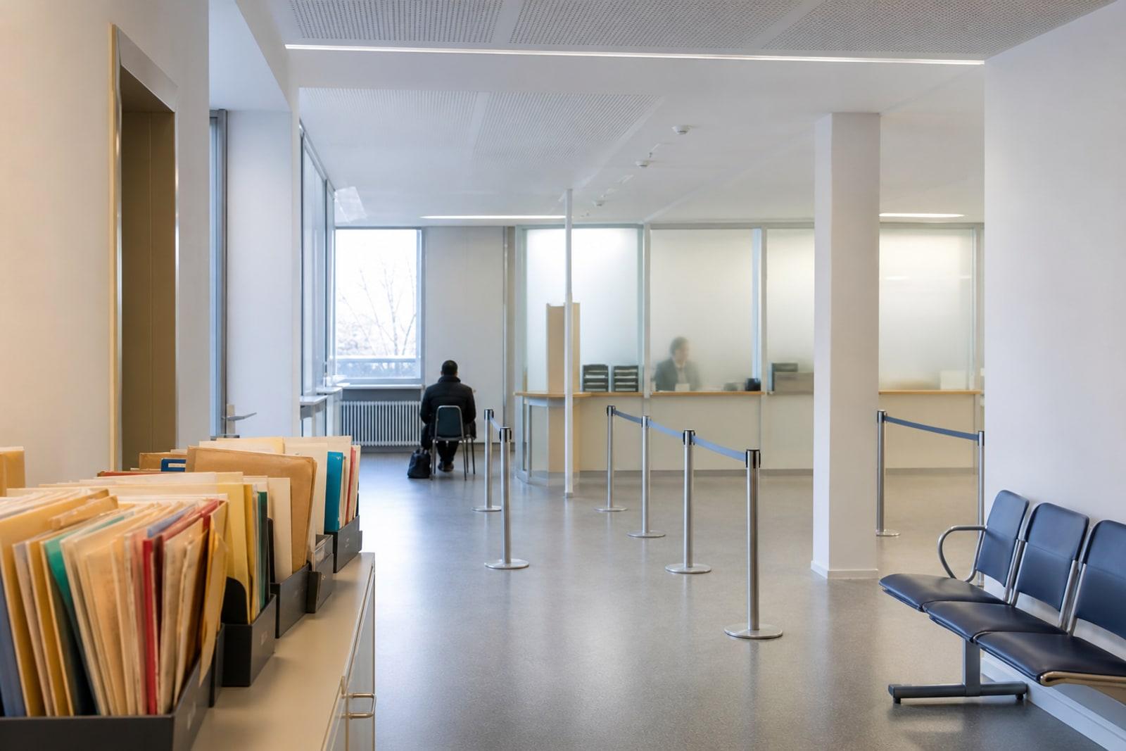 A quiet Luxembourg administrative waiting area with empty chairs, file folders, and anonymous distant figures near frosted service windows.