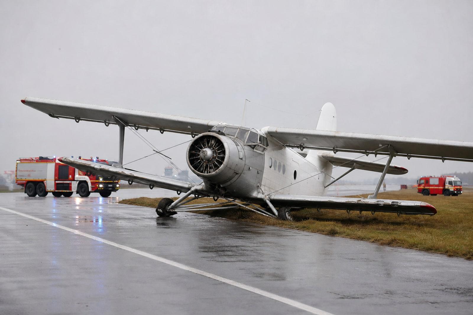 A vintage Antonov An-2 single-engine biplane stopped at the edge of an airport runway with a collapsed main landing-gear leg as fire-and-rescue vehicles stand by.