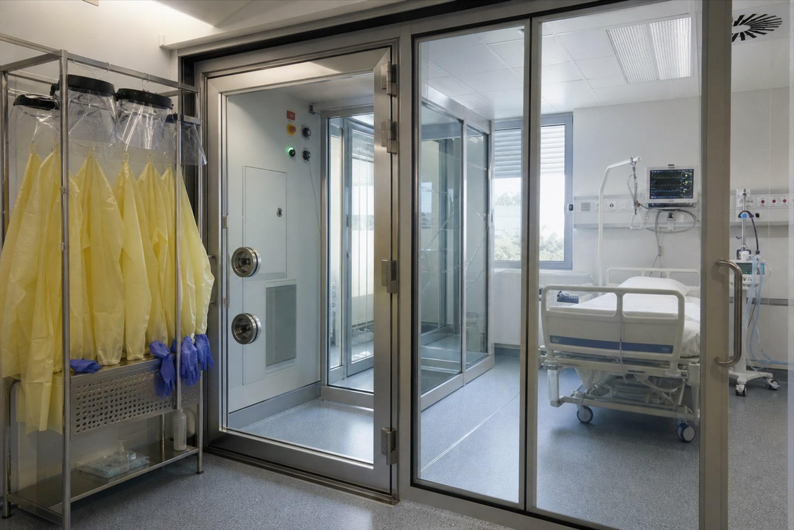 An empty hospital negative-pressure isolation room with a glass airlock and protective gowns and face shields on a rack.