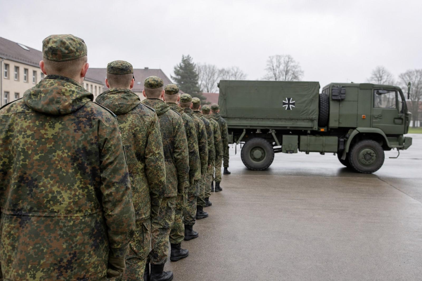 German Bundeswehr recruits in five-colour Flecktarn camouflage stand in line on a barracks parade ground beside a military vehicle bearing the black Iron Cross emblem.