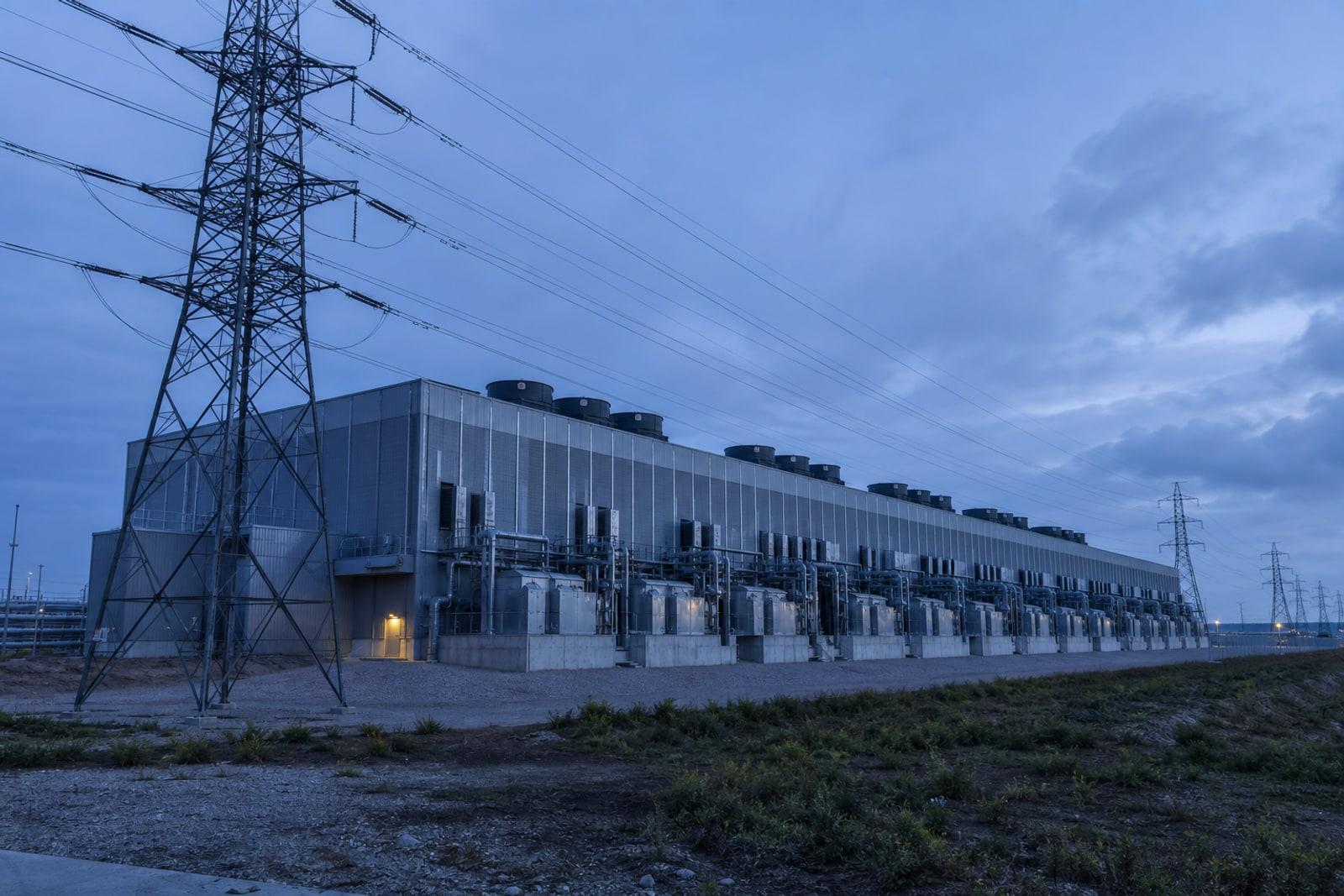 A large windowless data-centre building at dusk lined with cooling units and flanked by high-voltage transmission pylons and power lines.