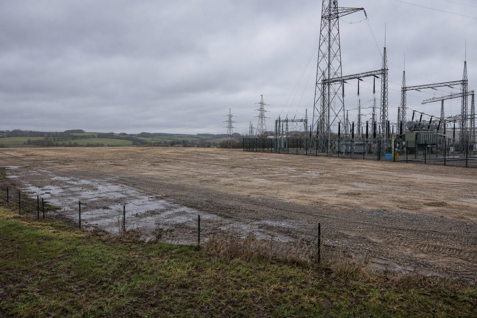 A flat cleared industrial plot at Bissen beside high-voltage pylons and a Creos electrical substation under an overcast sky.