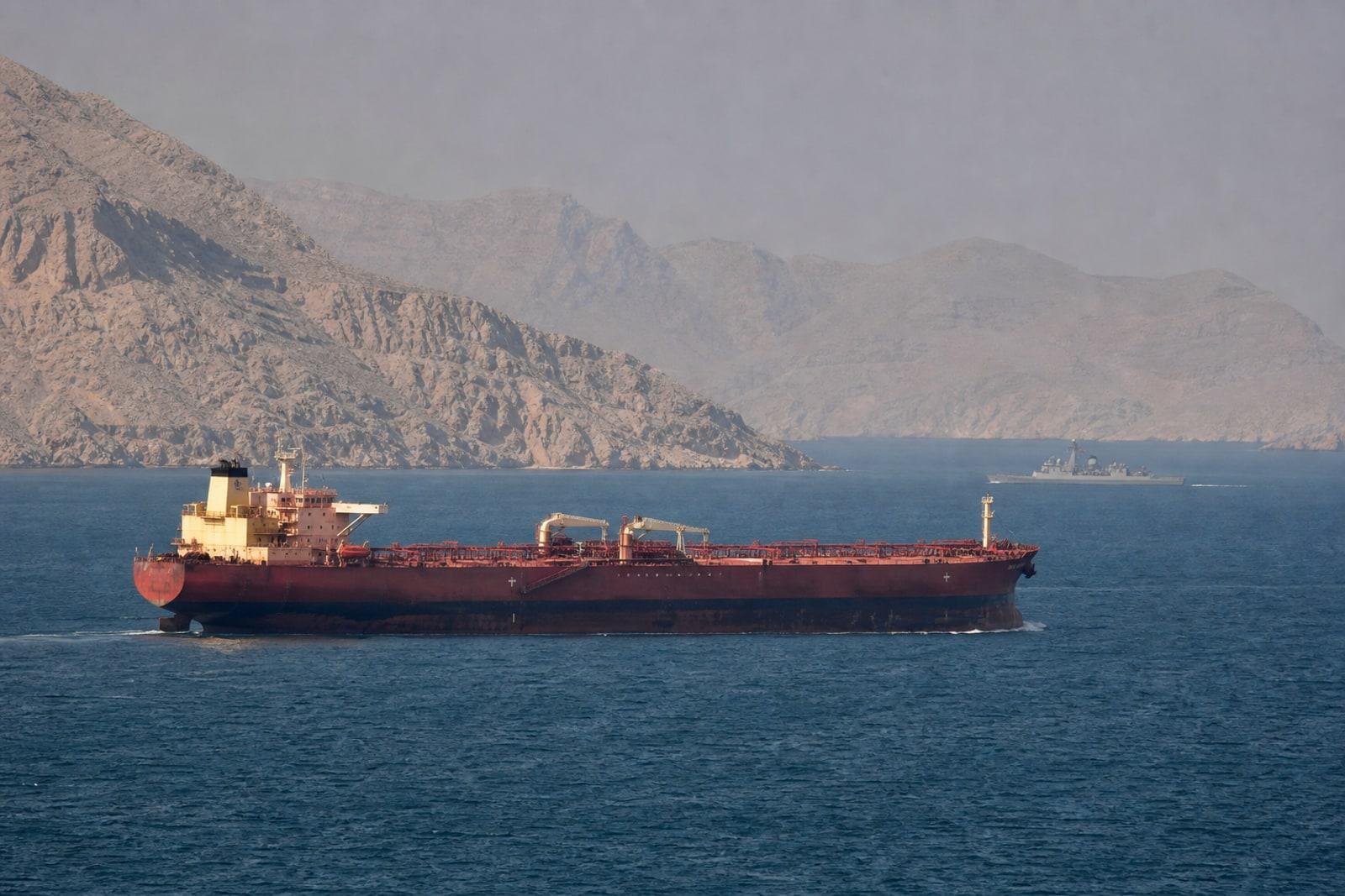 A laden crude-oil supertanker transits the narrow Strait of Hormuz between arid mountains, shadowed by a distant grey warship.