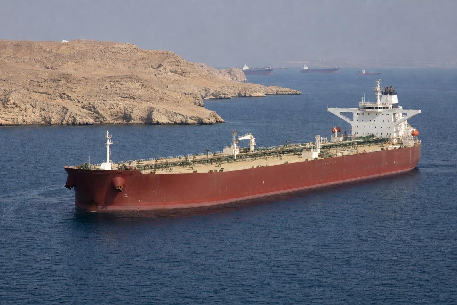 A laden crude oil supertanker transits the Strait of Hormuz past an arid coastline under clear skies.