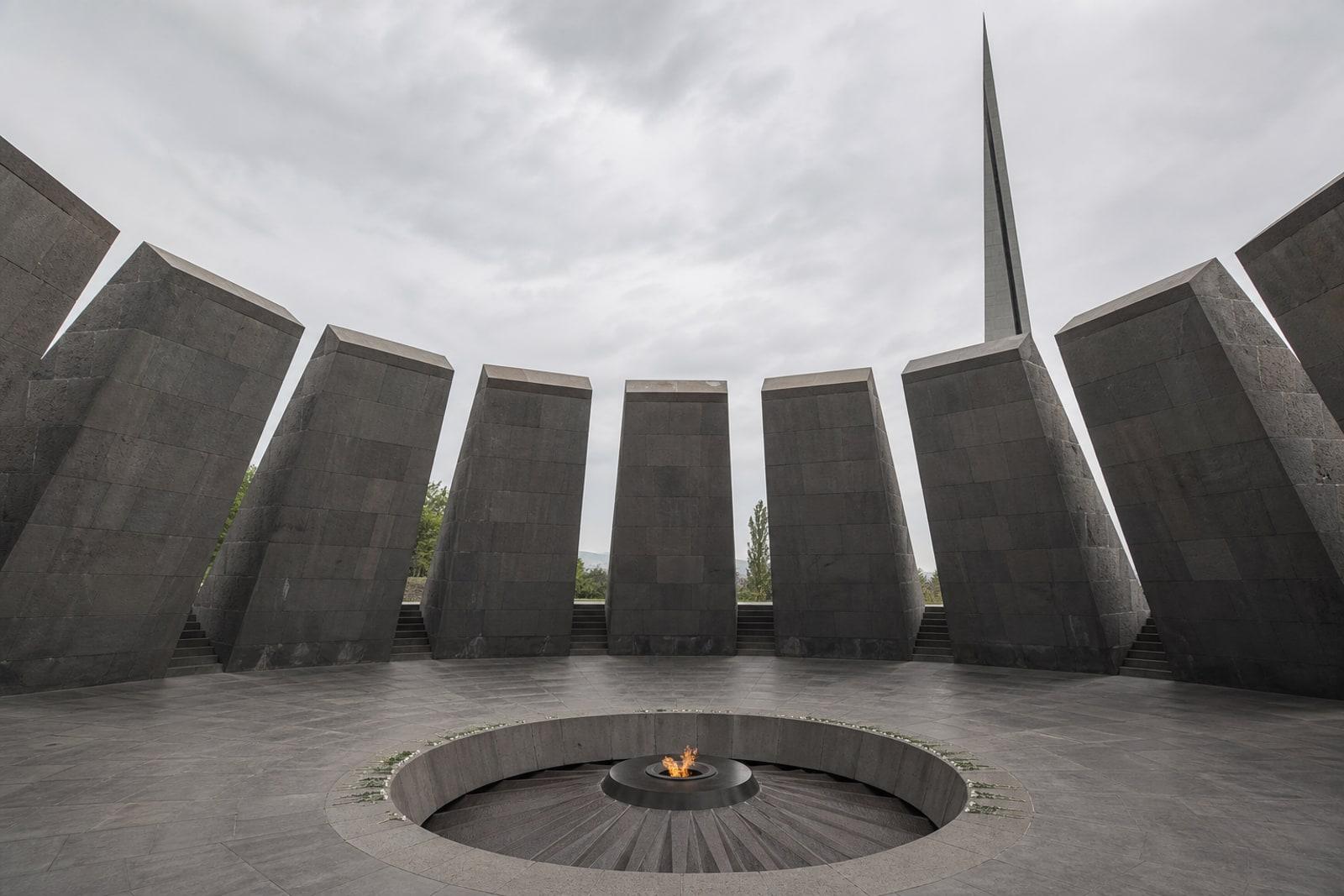 The eternal flame within a circle of twelve tall leaning basalt slabs at the Armenian Genocide memorial, with a tall split stele behind under grey sky.
