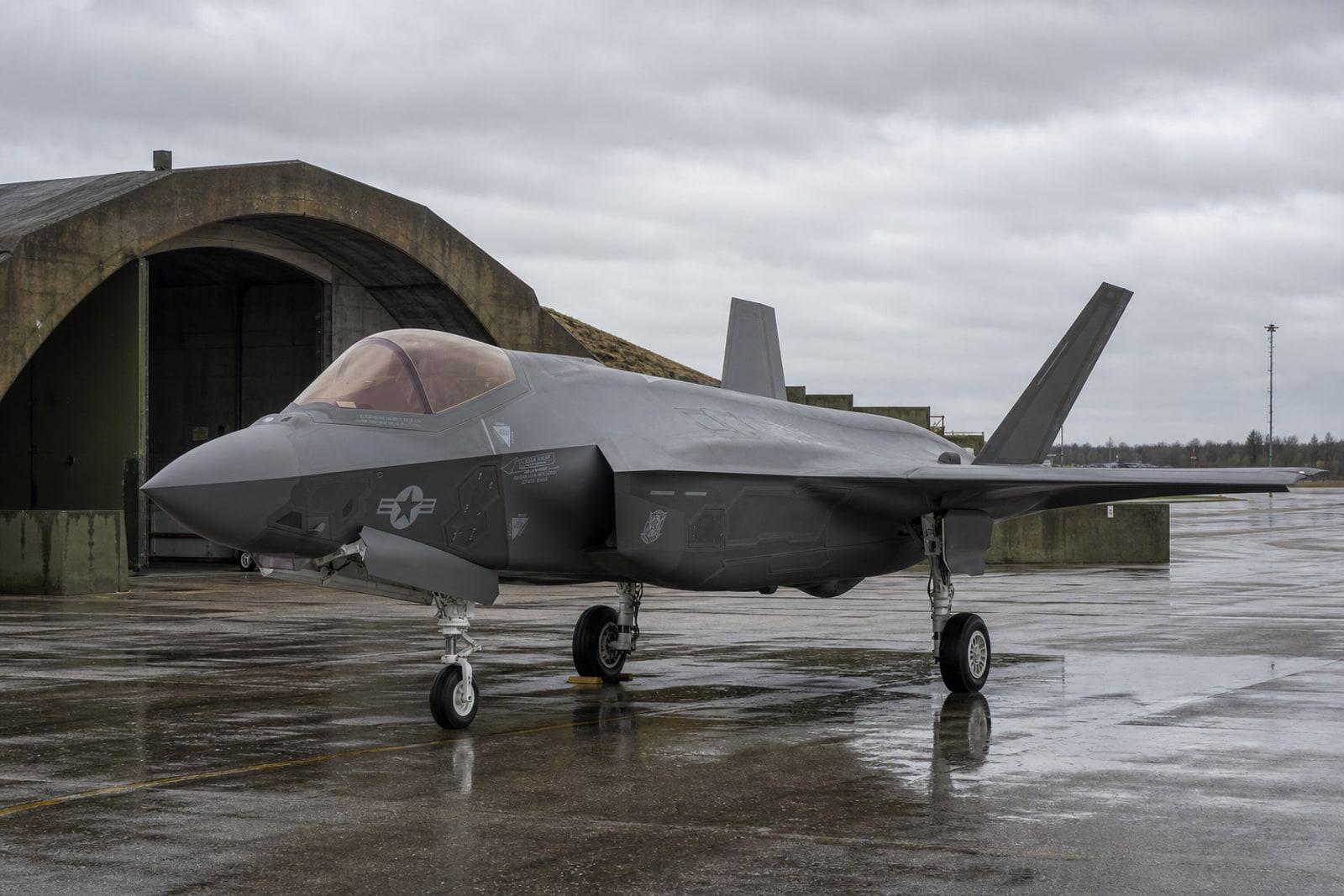 A matte-grey F-35A Lightning II stealth fighter parked on a wet airbase apron under an overcast Baltic sky.