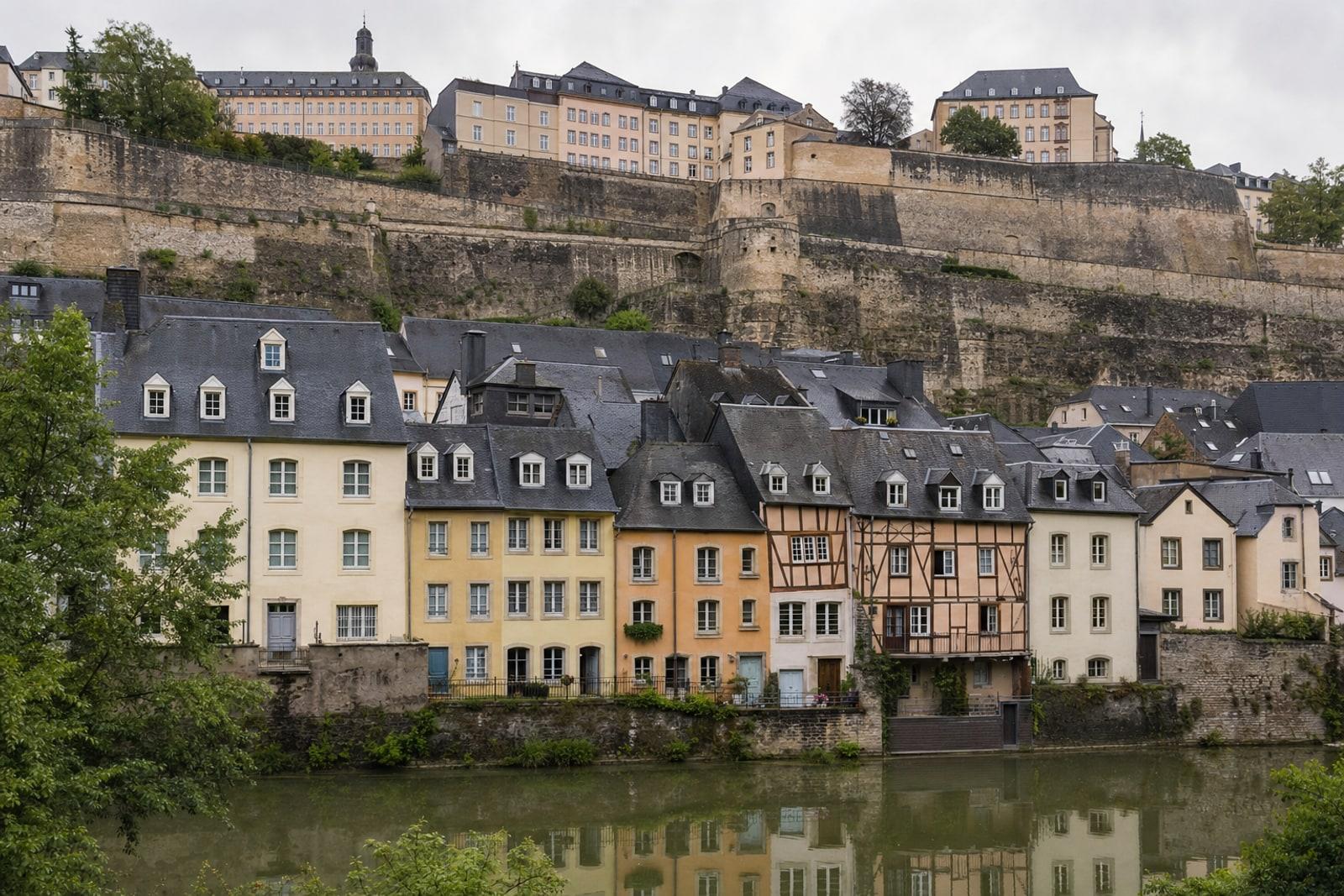 Pastellfaarweg Haiser mat Schifferdiech am Gronn laanscht d'Uelzecht, driwwer d'Festungsmaueren vun der Aler Stad.