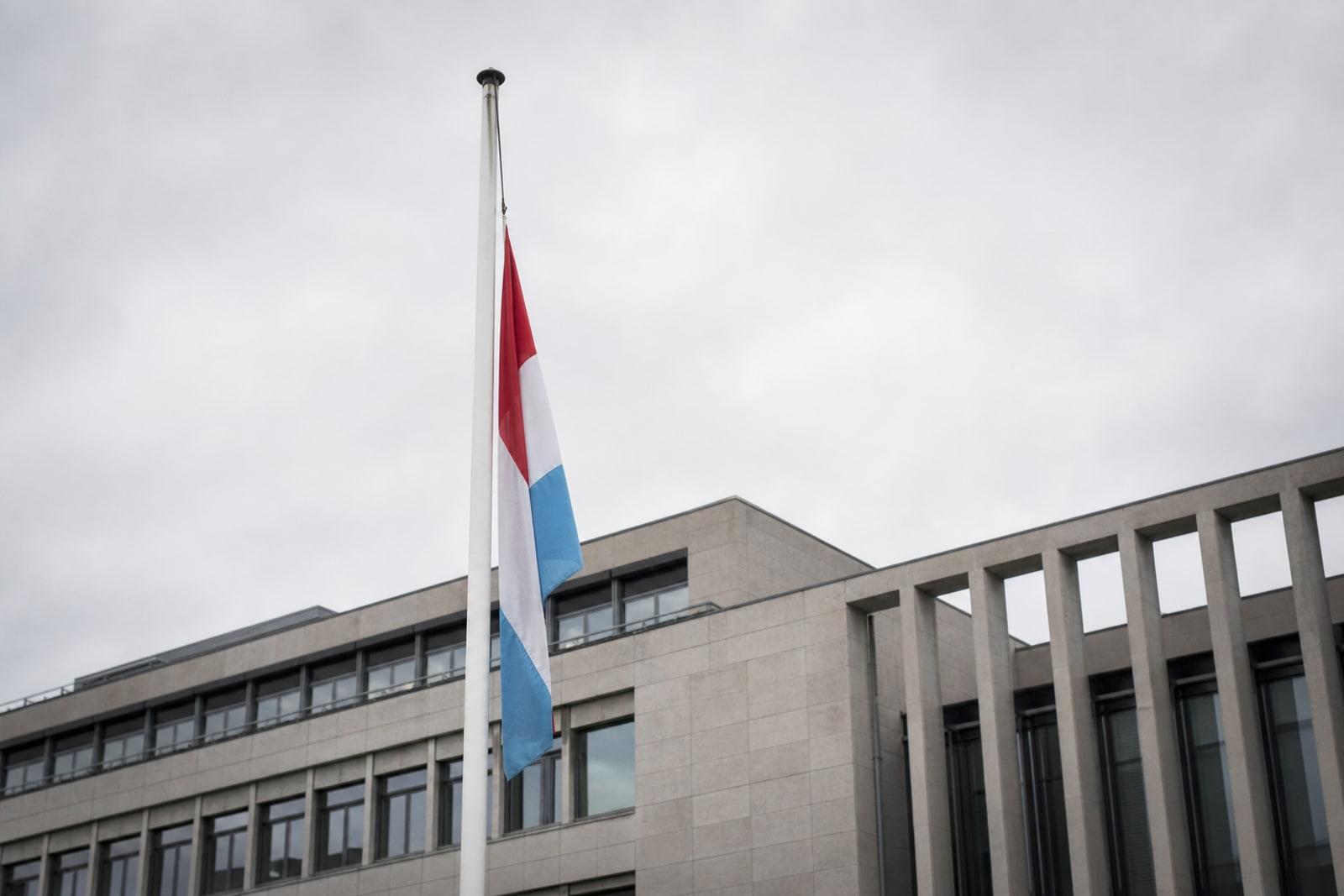 The national flag of Luxembourg flying on a pole outside a modern government building under an overcast sky.