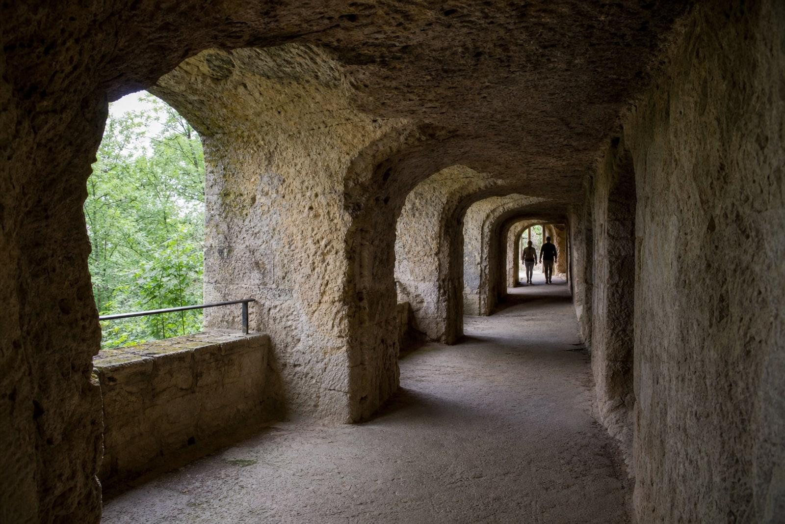 A quiet Luxembourg fortress casemate tunnel with rough stone arches, summer daylight, greenery outside, and two anonymous distant visitors walking away.