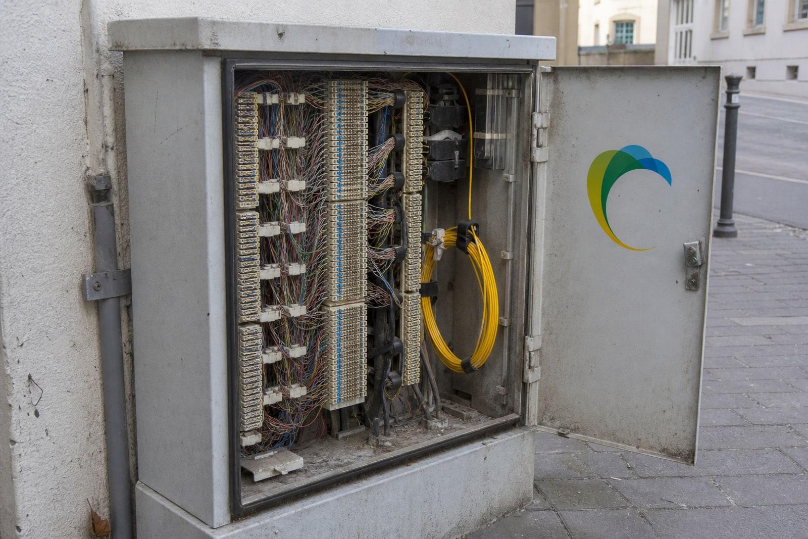 An open POST Luxembourg street cabinet showing bundles of copper telephone wiring alongside a coil of yellow fibre.