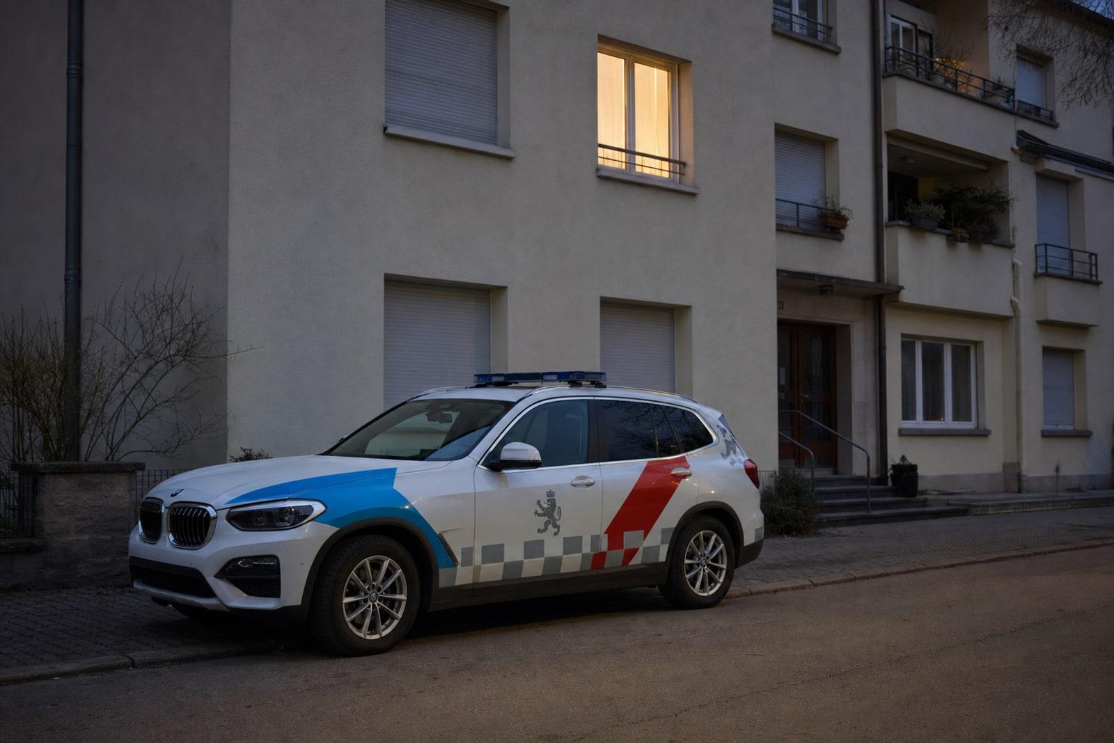 A white Luxembourg police car with blue-and-red markings parked at dusk outside a residential apartment building.