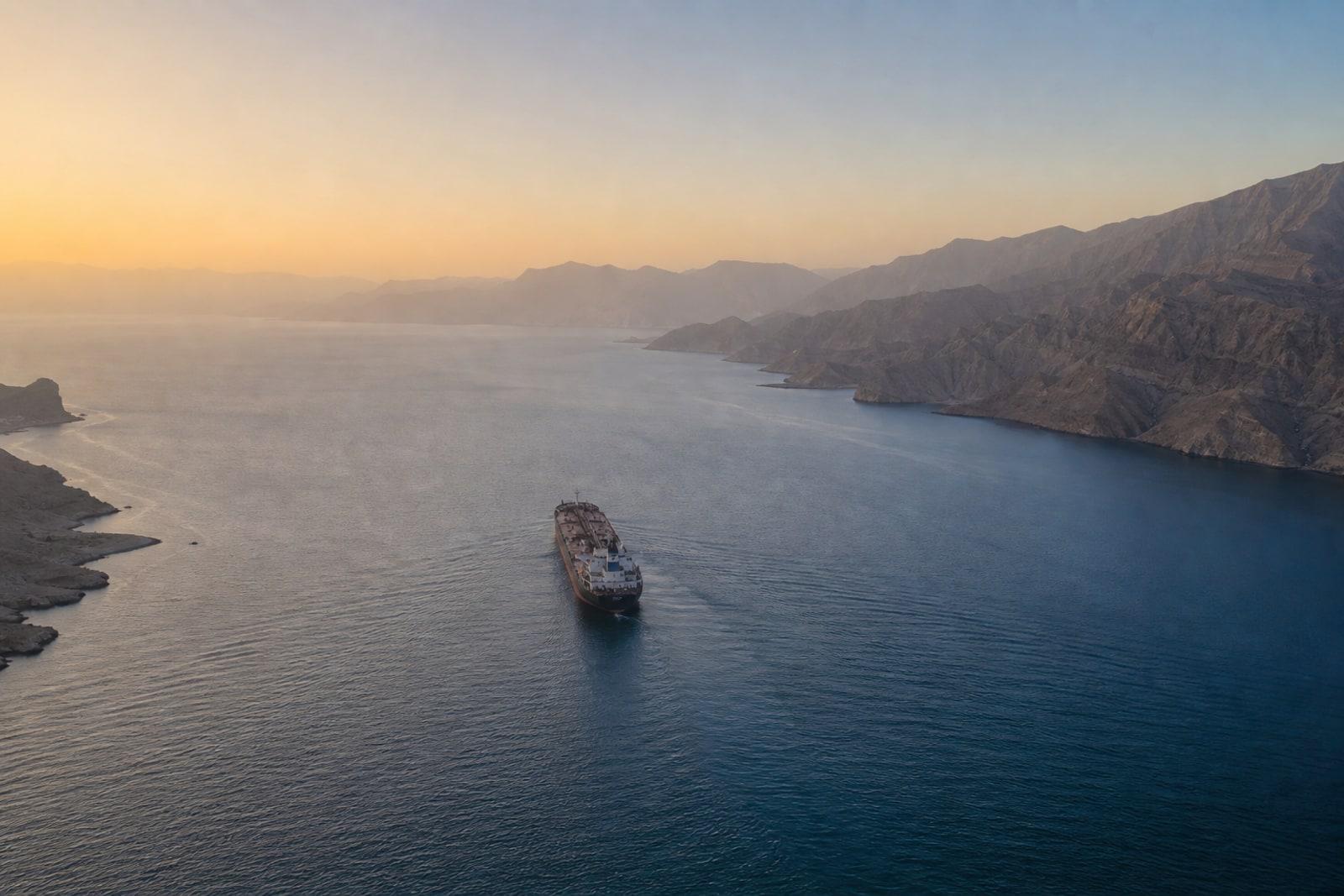 A distant oil tanker moving through the narrow waters of the Strait of Hormuz at dawn, framed by hazy coastal mountains.