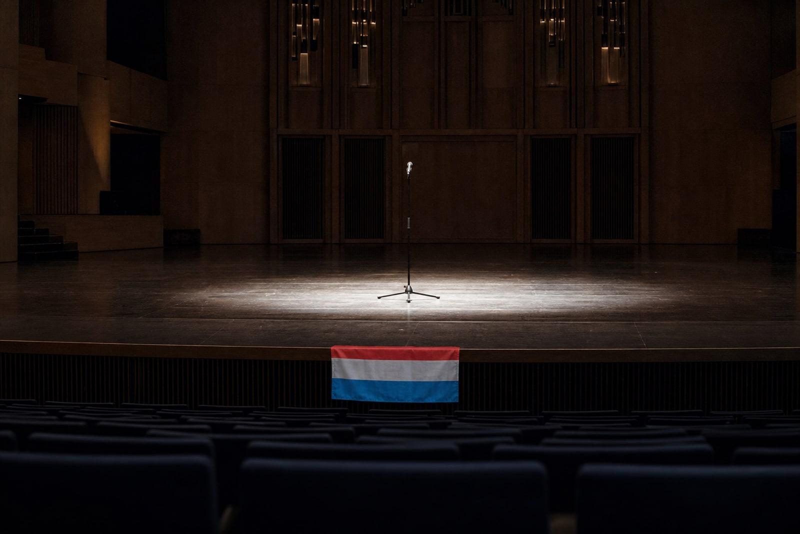 An empty national-final stage in a concert hall lit by a single spotlight on a bare microphone stand with a small Luxembourg flag.