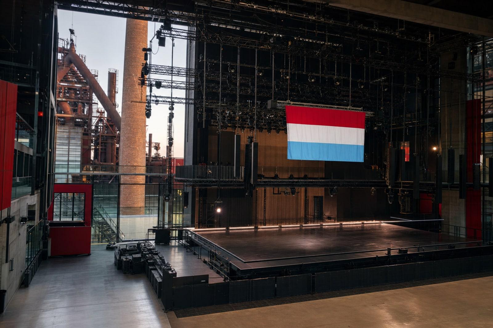 An empty, brightly lit contest stage inside a modern concrete-and-steel concert hall, with a large Luxembourg red-white-and-light-blue flag hanging above it.