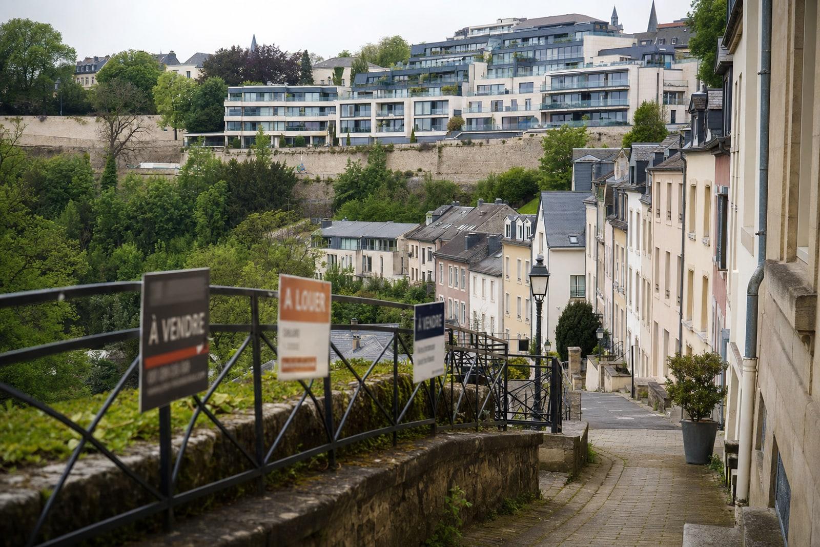 A terraced residential street in Luxembourg City with townhouses and apartment blocks and estate-agent for-sale and to-let signs.