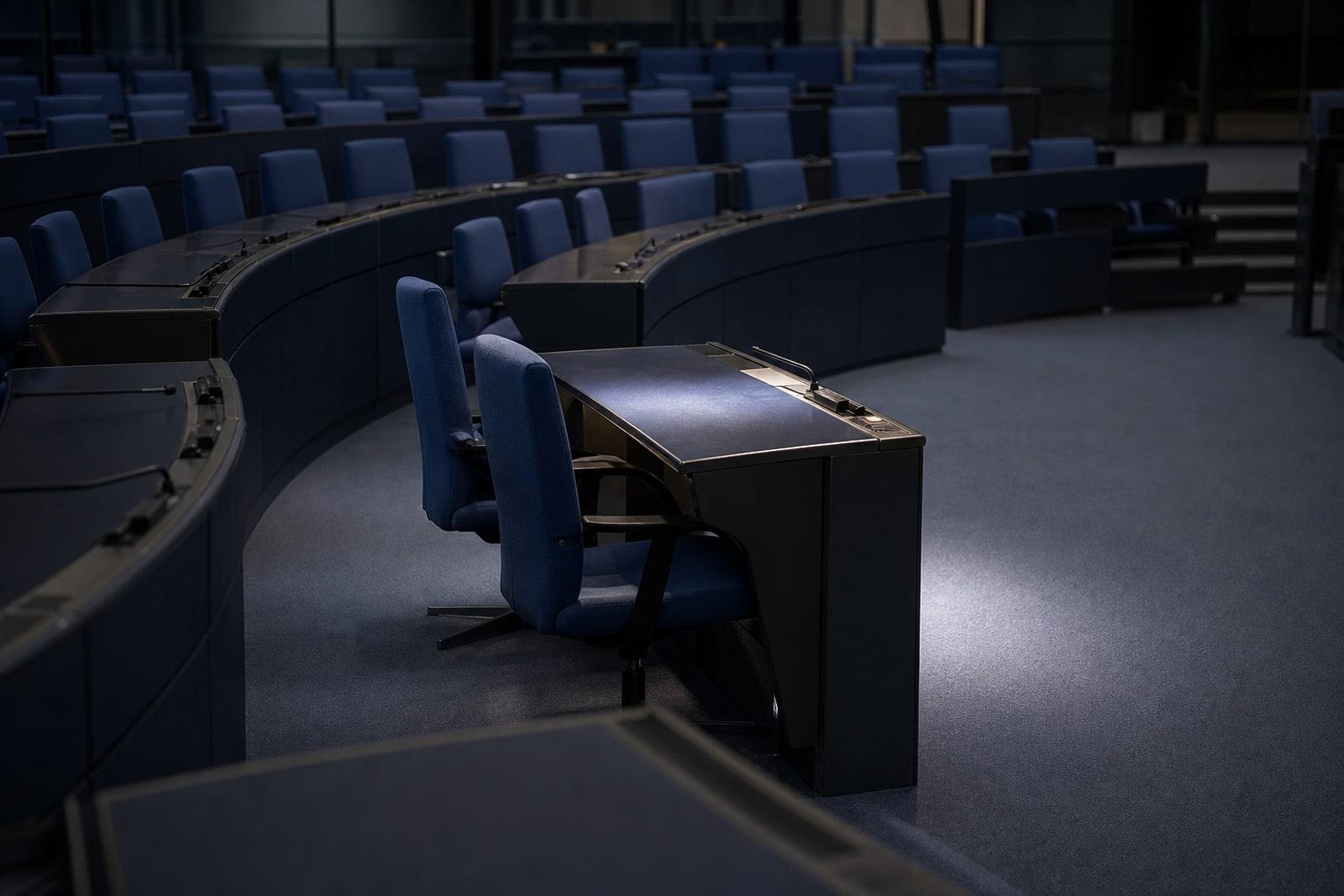 A single empty desk and seat among the curved blue benches of a vast, quiet parliamentary chamber.