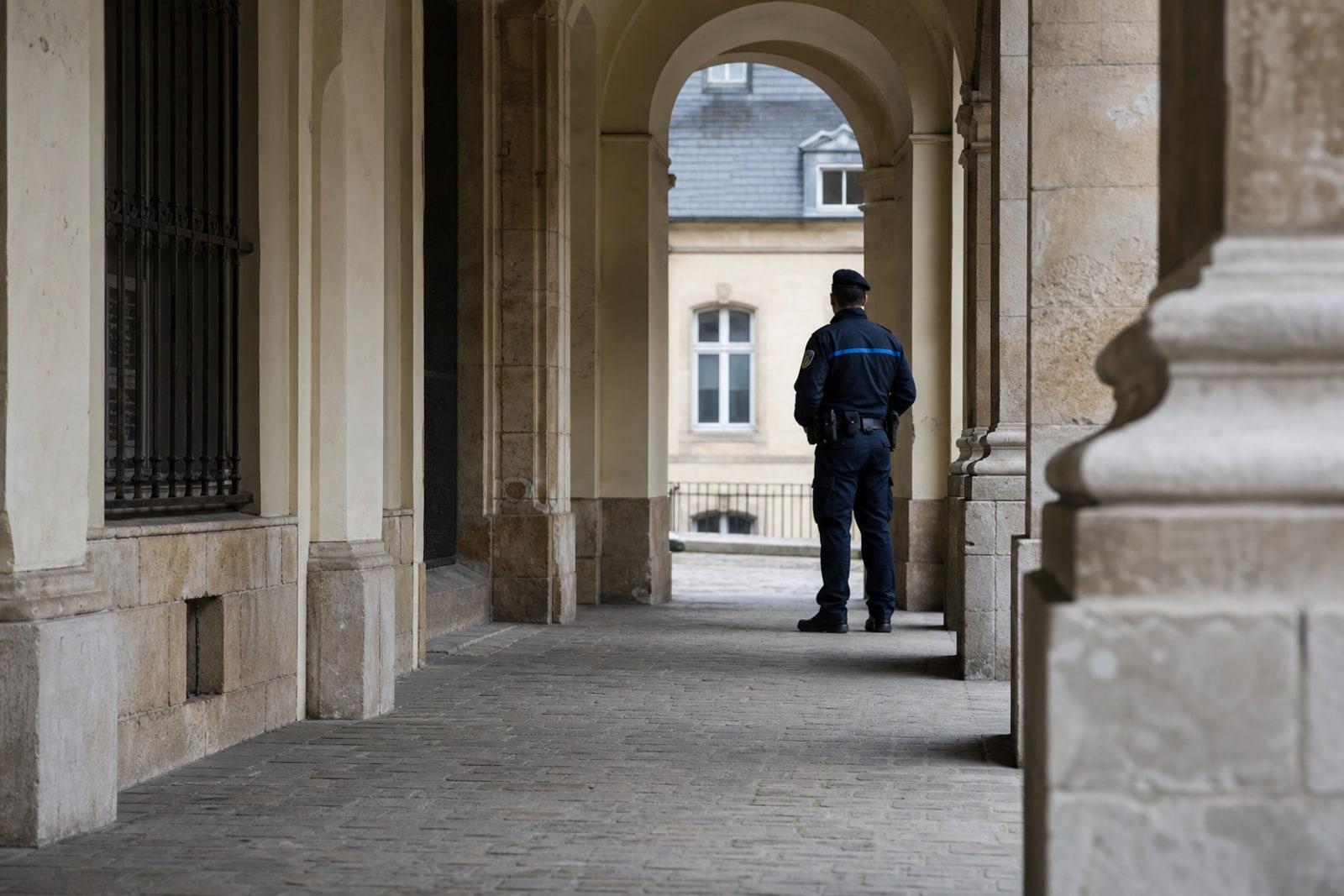 A Grand Ducal Police officer in dark navy-blue uniform standing in an empty arcaded stone street in Luxembourg City's old town.