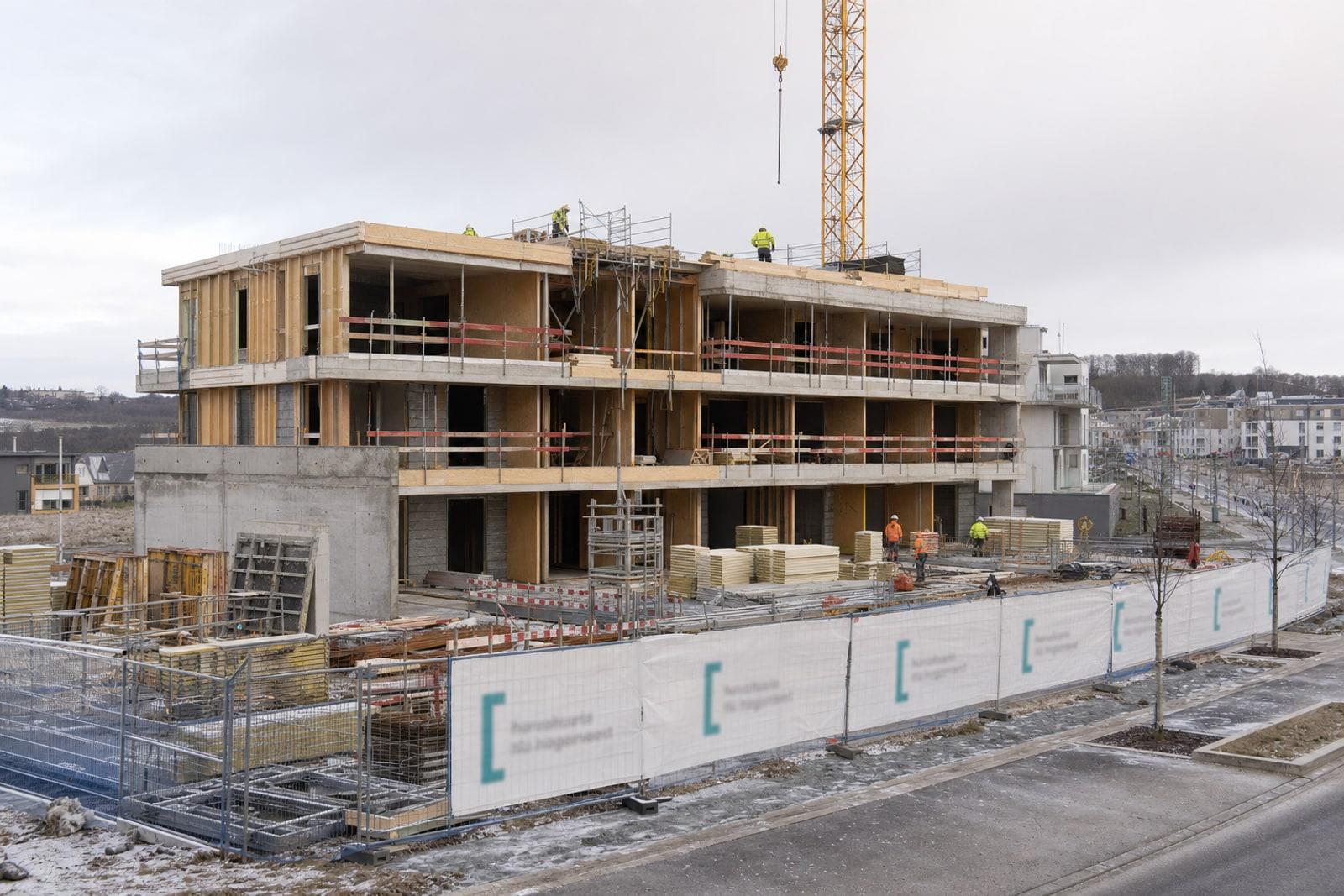 An affordable-housing apartment block under construction in Luxembourg, with scaffolding, a tower crane and workers on the concrete frame.