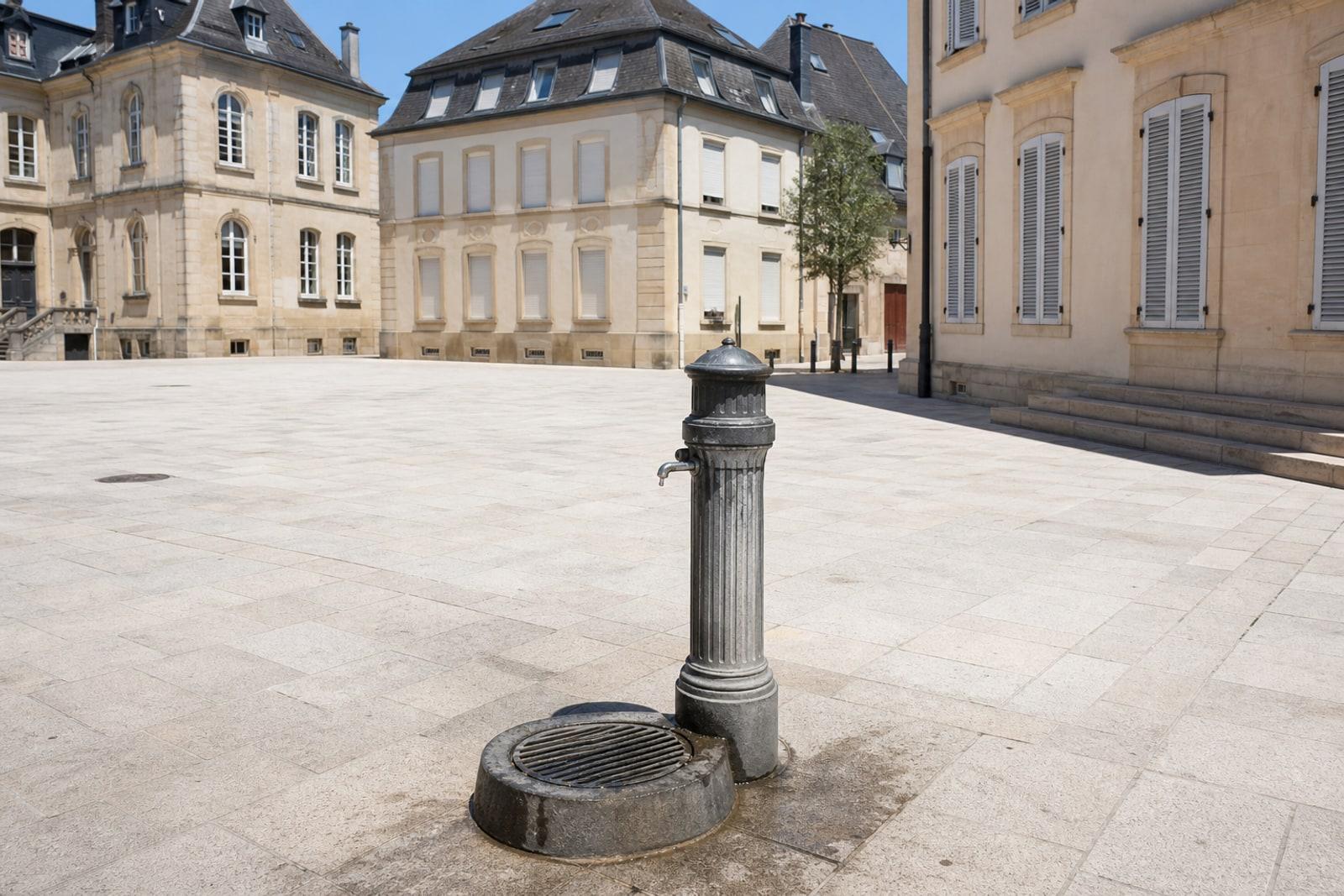 A public drinking-water fountain on an empty, sunlit city square in Luxembourg under a harsh midday sun.