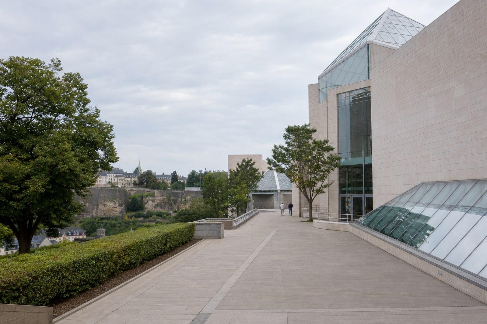 A quiet Luxembourg institutional terrace beside a pale stone and glass EU-style administrative building, with two anonymous figures walking in the distance under overcast daylight.