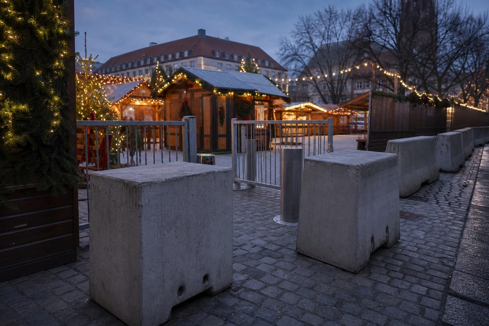 Gesicherter Zugang zu einem europäischen Weihnachtsmarkt unter freiem Himmel: graue Beton-Sperrblöcke und ein kontrolliertes Eingangstor vor beleuchteten Holzbuden in der Abenddämmerung.