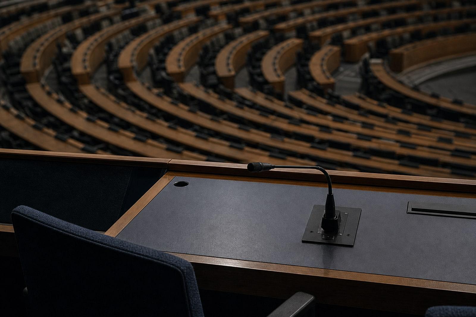An empty seat and switched-off microphone in a dim, deserted parliamentary debating chamber seen from above.