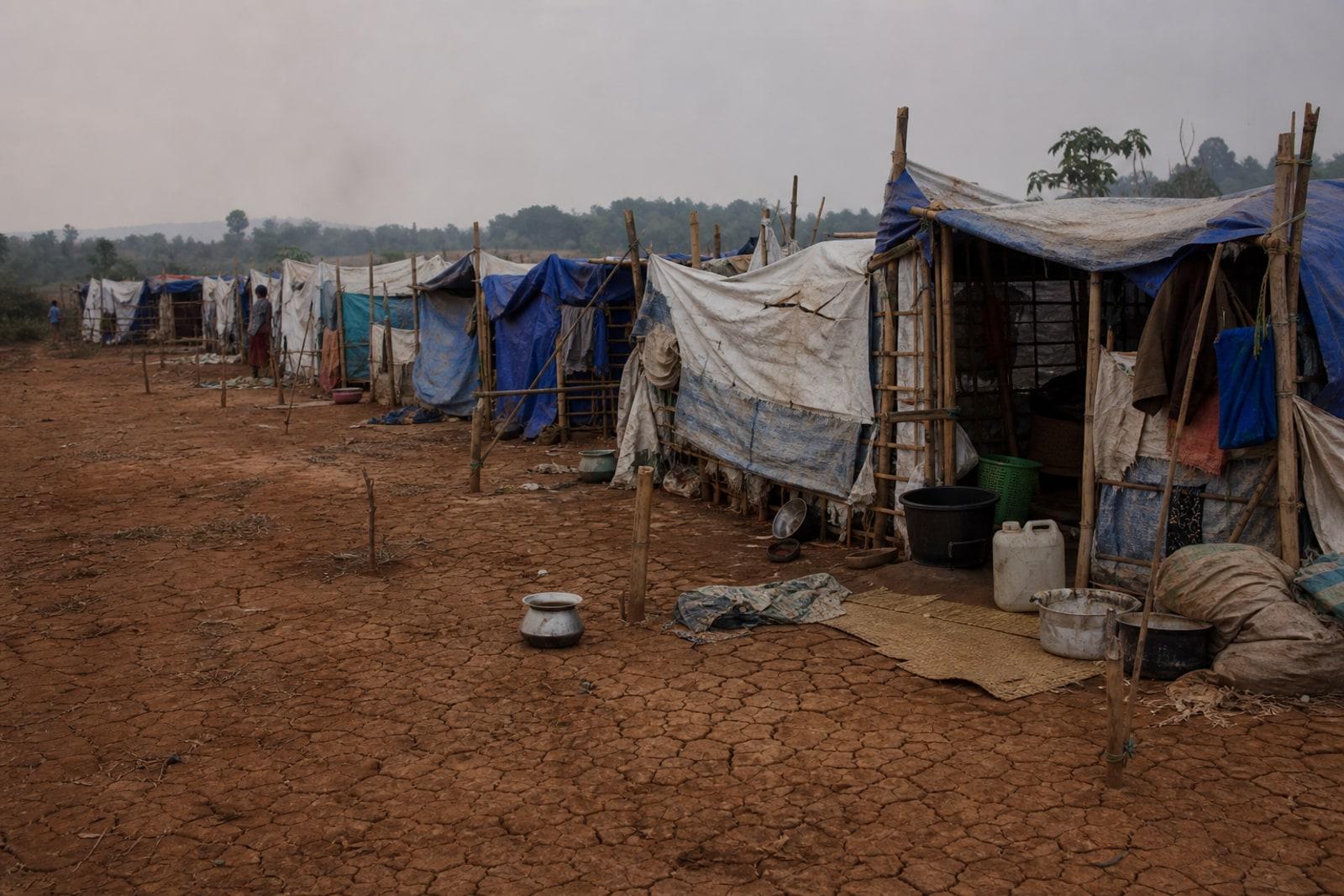 Rows of bamboo-and-tarpaulin shelters at a makeshift camp for internally displaced people on cracked earth under a hazy sky in central Myanmar.