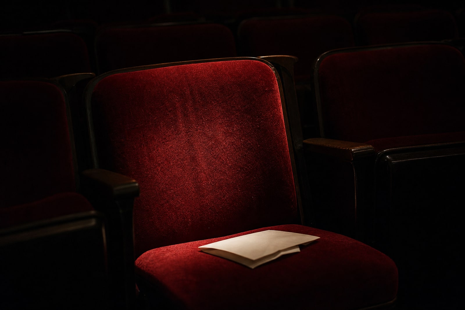 An empty illuminated seat in a darkened classical concert hall with a folded programme left on it.