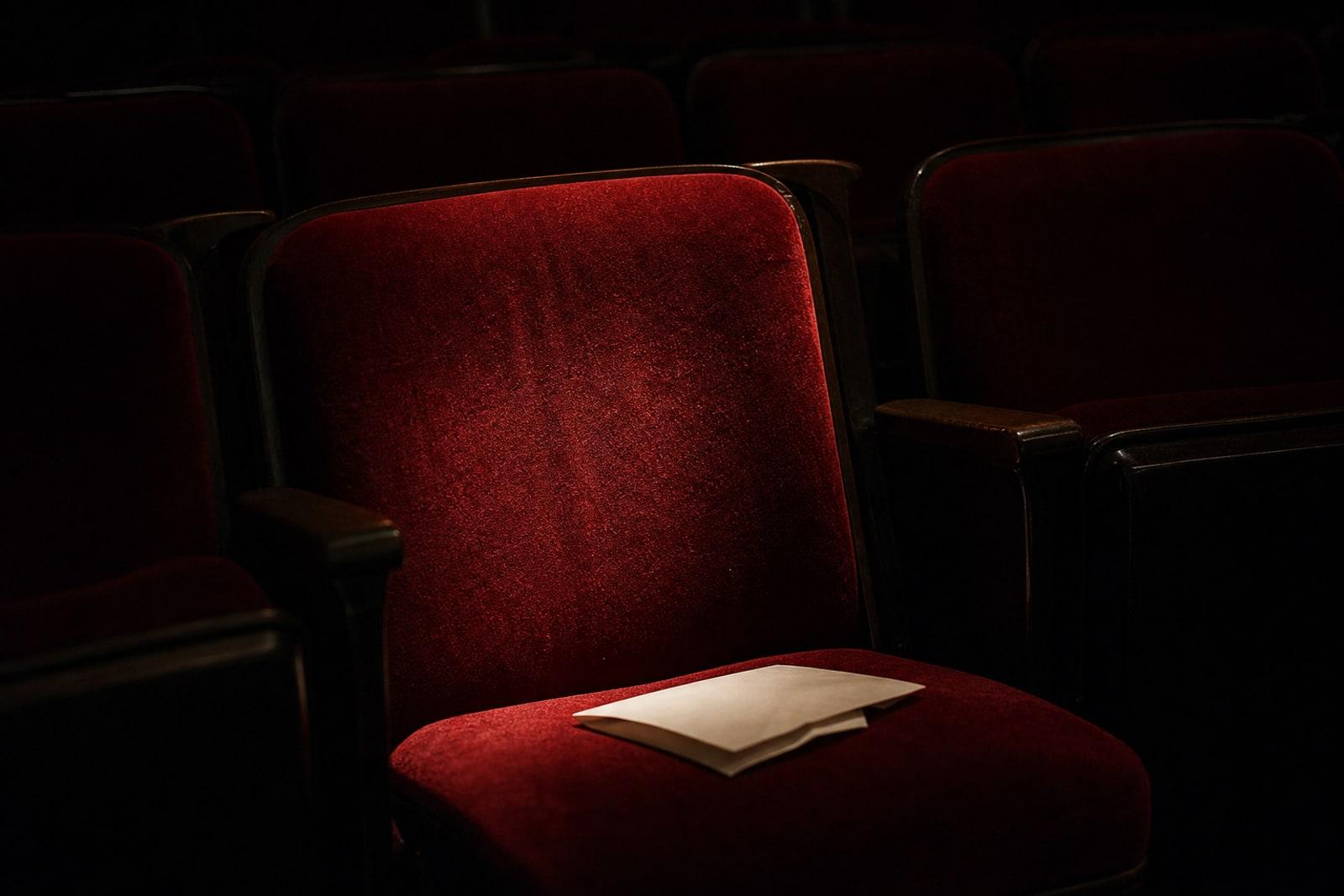 An empty illuminated seat in a darkened classical concert hall with a folded programme left on it.