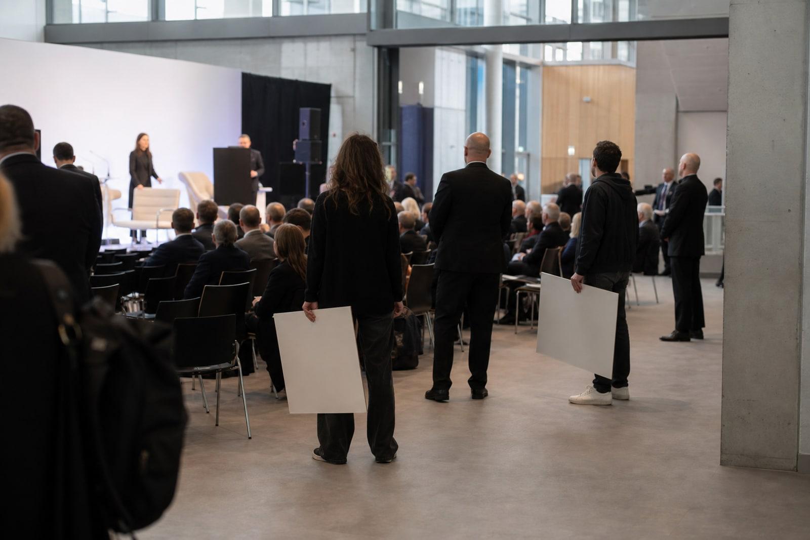 Anonymous protesters with blank placards stand at the back of a modern conference hall during a distant tech summit presentation.