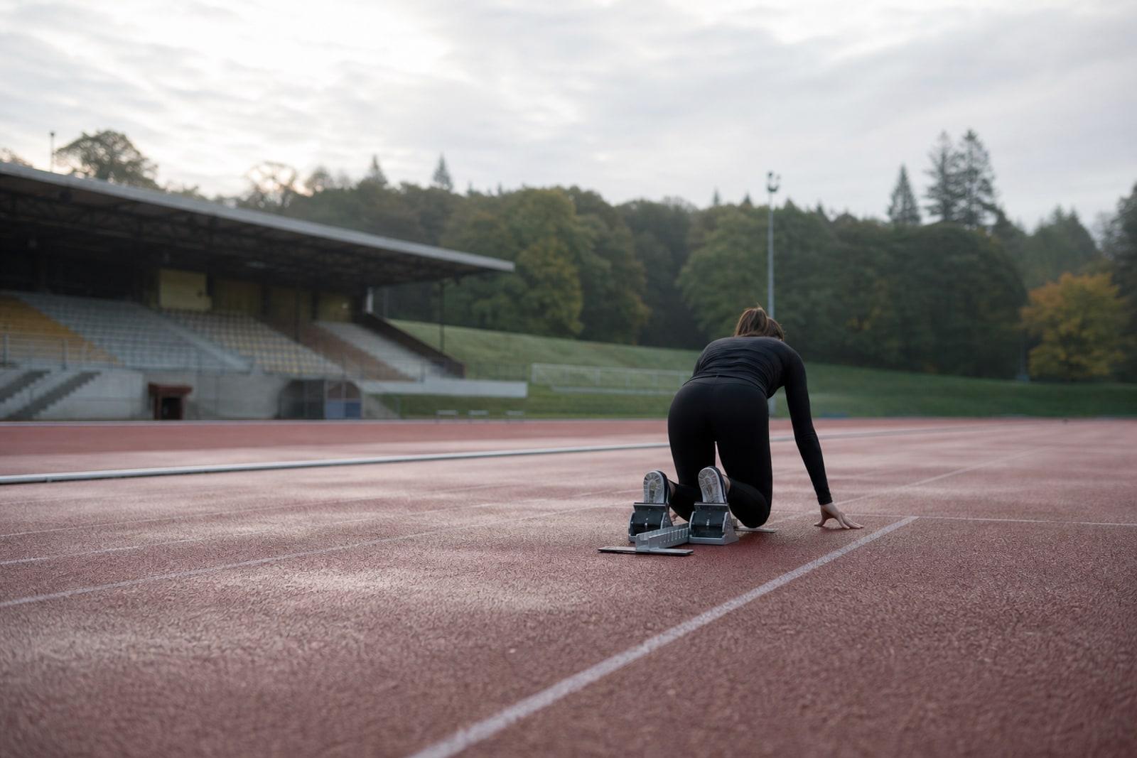 Anonymous sprinter seen from behind in starting blocks on a damp outdoor track beside a quiet stadium and tree line.