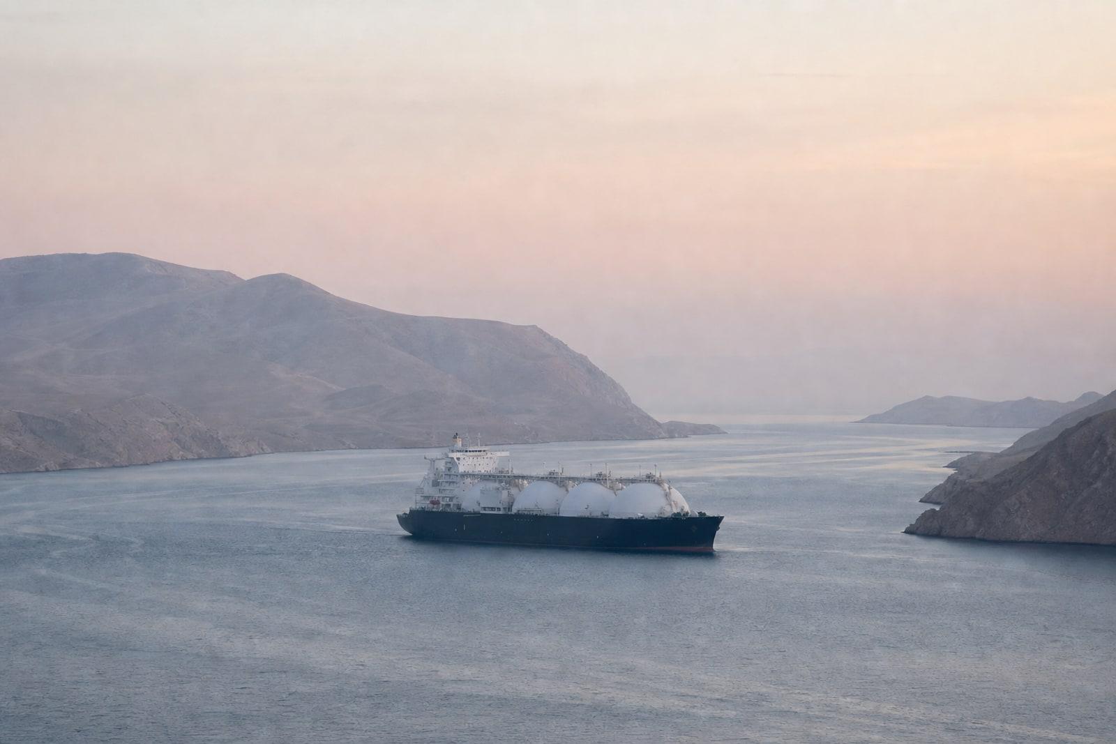 An LNG carrier with spherical tanks transits a narrow sea strait between bare desert mountains in dawn light.