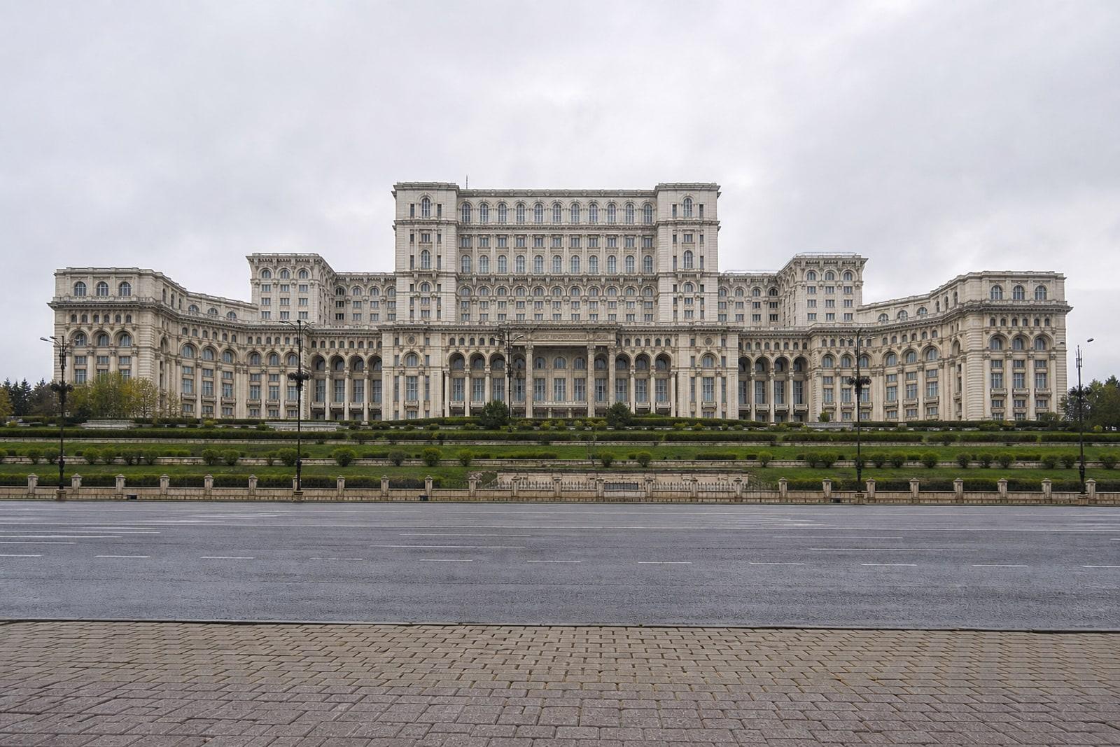 The vast white limestone Palace of the Parliament in Bucharest under an overcast sky, seen from the empty boulevard before it.