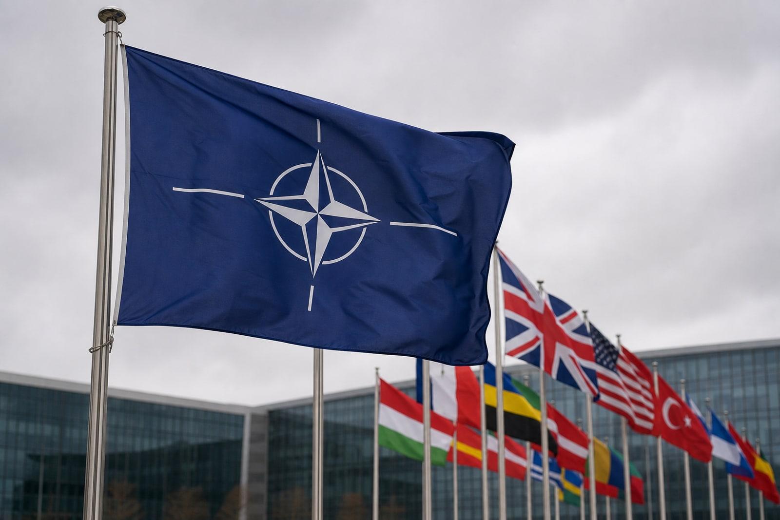 The blue NATO flag with its white compass-star emblem flying beside allied national flags outside a summit building.