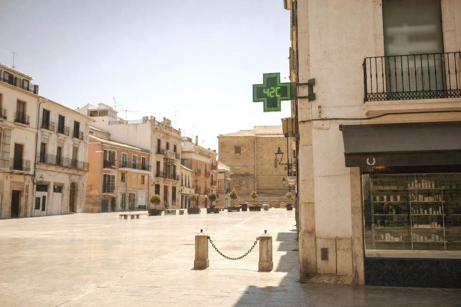 A near-empty sun-bleached Spanish city plaza at midday, with a green pharmacy cross displaying a high temperature under a hazy sky.