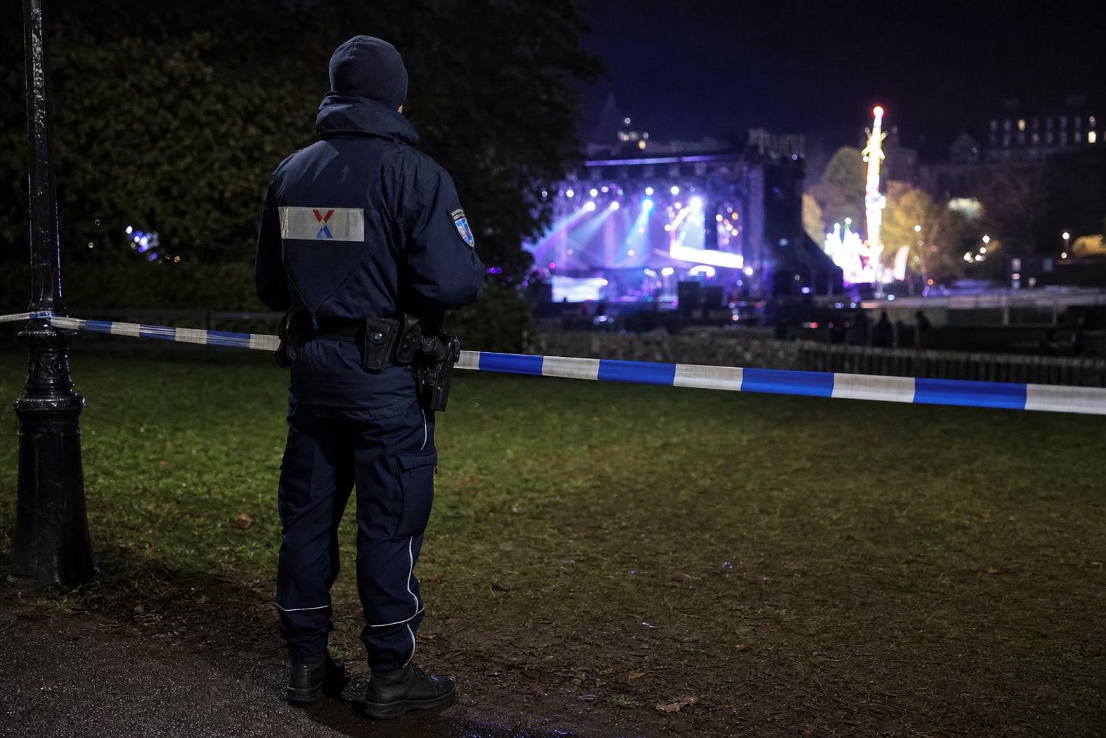 A navy-uniformed Grand Ducal Police officer stands beside blue-and-white police tape at night on the grassy edge of Luxembourg City's Champ du Glacis, with blurred concert and fairground lights behind.