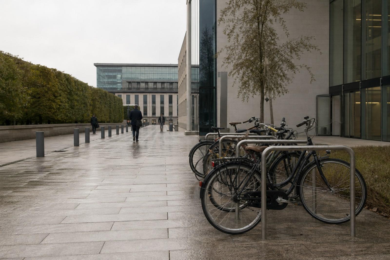 Damp paving, parked bicycles and distant anonymous commuters outside a modern Luxembourg civic office building in soft morning light.